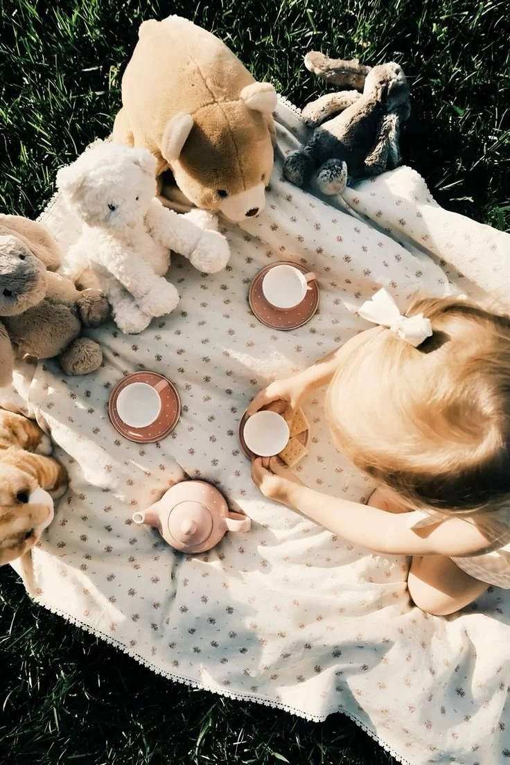 Child having a tea party outdoors on a blanket with stuffed animals and miniature tea set.