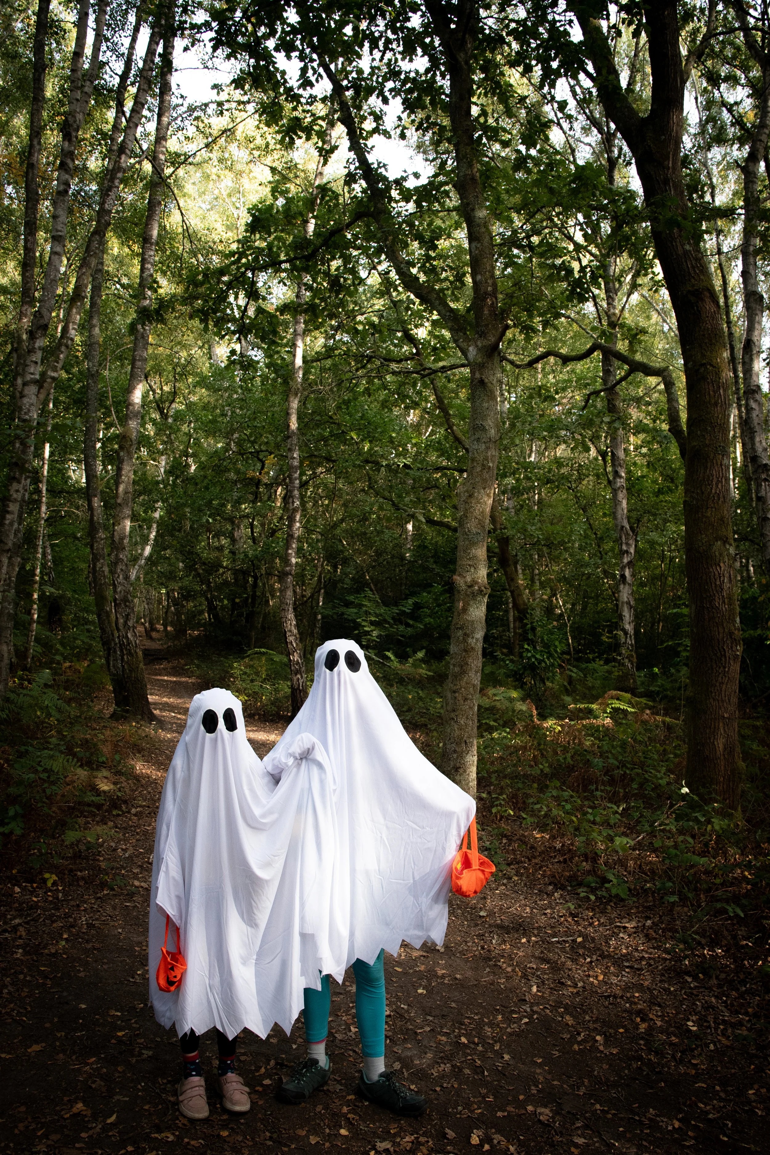 Two children dressed in ghost costumes made of white sheets with eye holes, standing on a forest trail surrounded by trees.