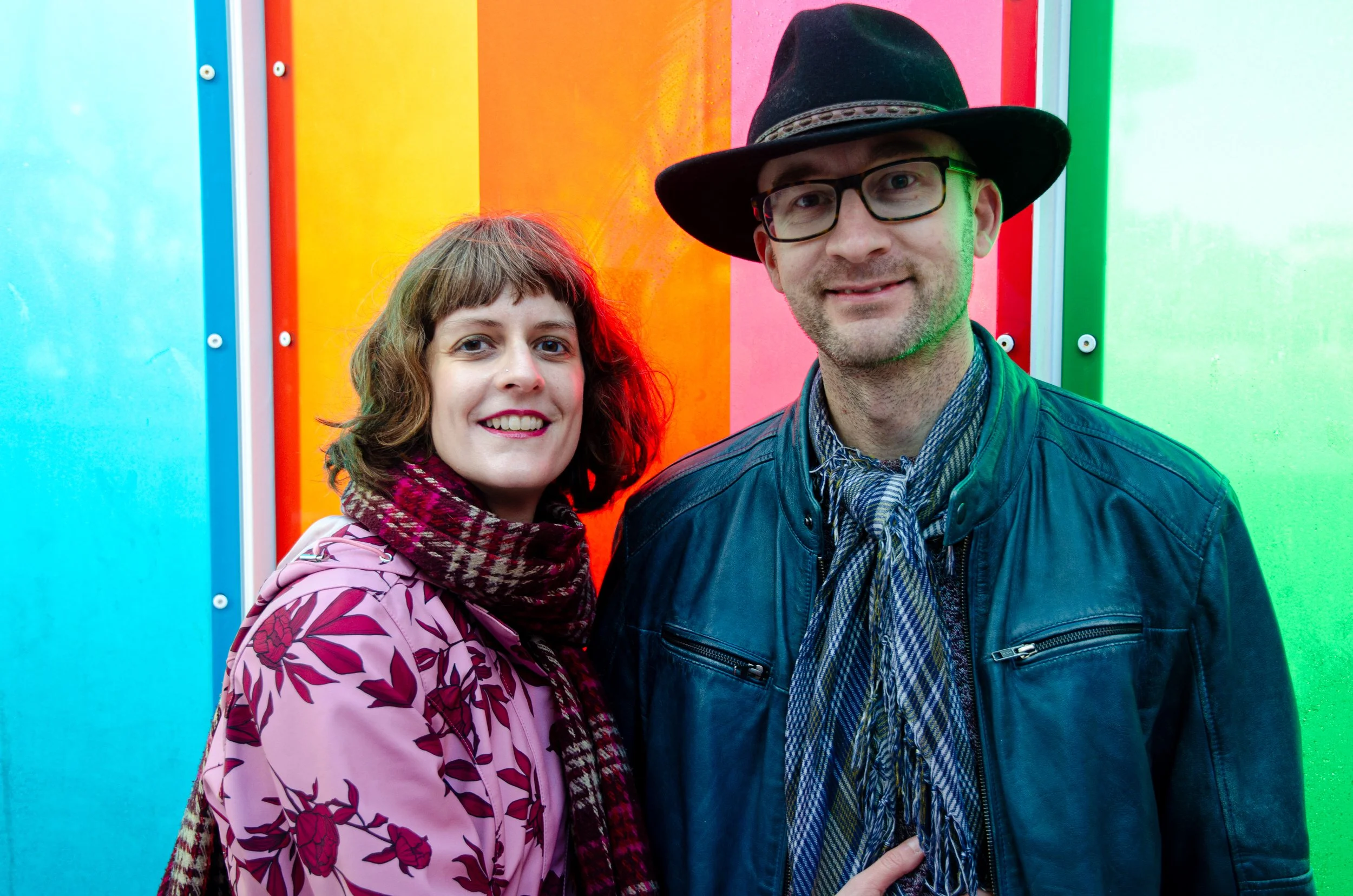 A man and woman smiling at the camera, standing in front of a colorful rainbow wall. The woman has shoulder-length brown hair and is wearing a pink jacket with red floral pattern and a matching scarf. The man has glasses and a beard, wearing a black wide-brimmed hat and a leather jacket with a plaid shirt underneath.