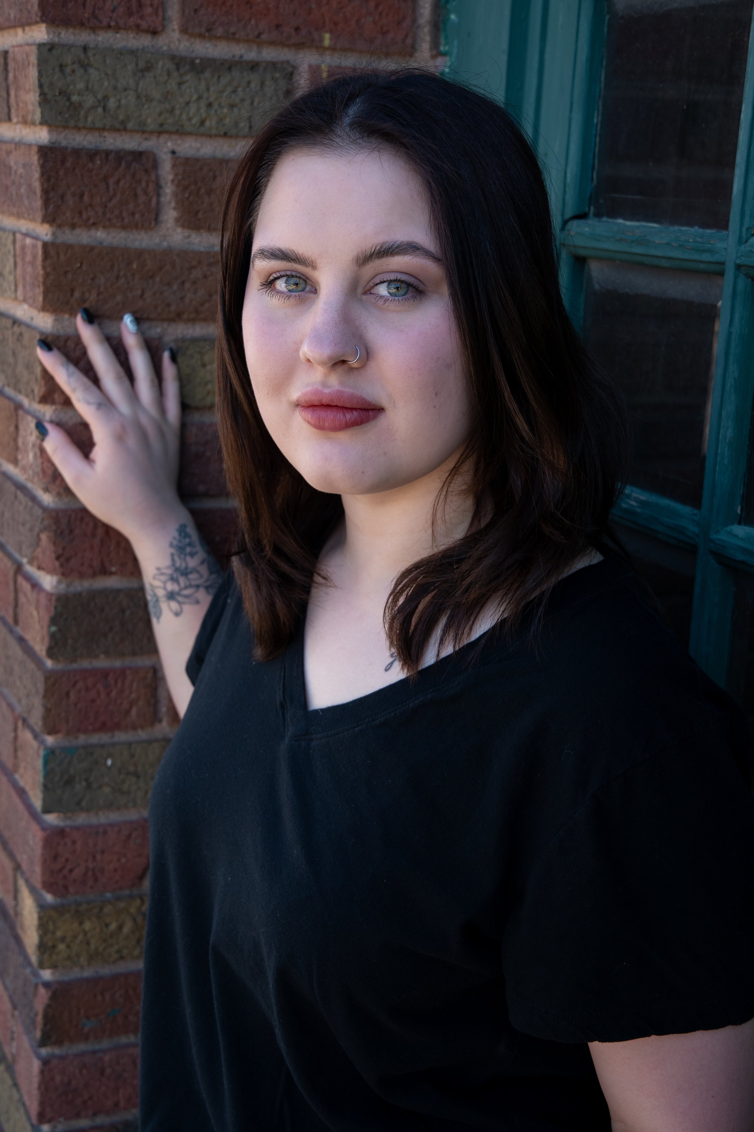 A woman with dark brown hair, fair skin, and blue eyes, wearing a black shirt, standing outdoors next to a brick wall and green window frame.
