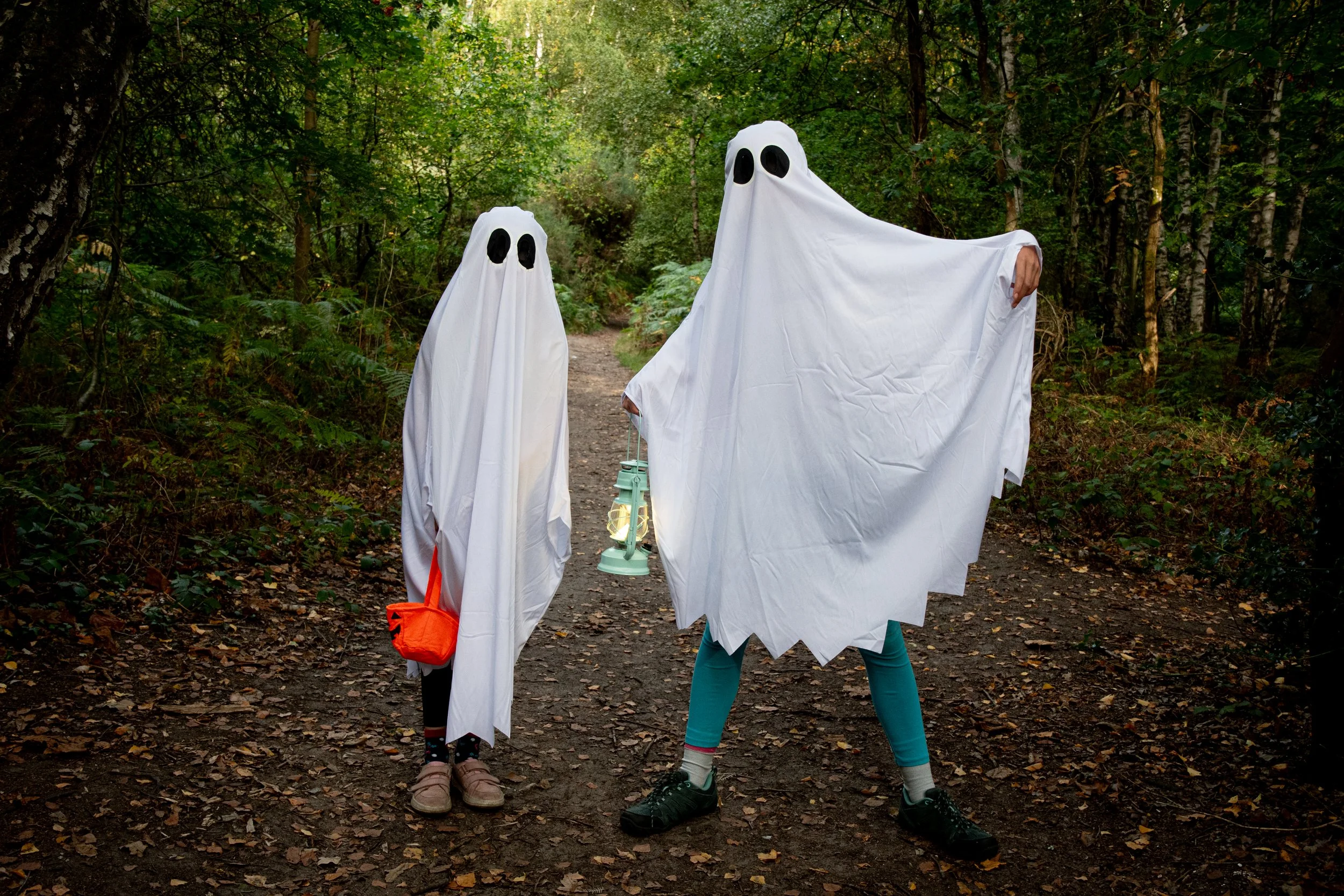 Two people dressed as ghosts with white sheets and black eye holes walk through a forested path.