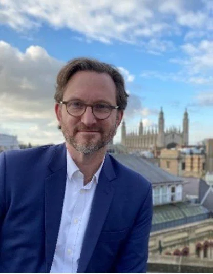 Man with glasses and beard in a blue suit and white shirt, standing outdoors with historic buildings and the cityscape of London, including Big Ben, in the background under a partly cloudy sky.