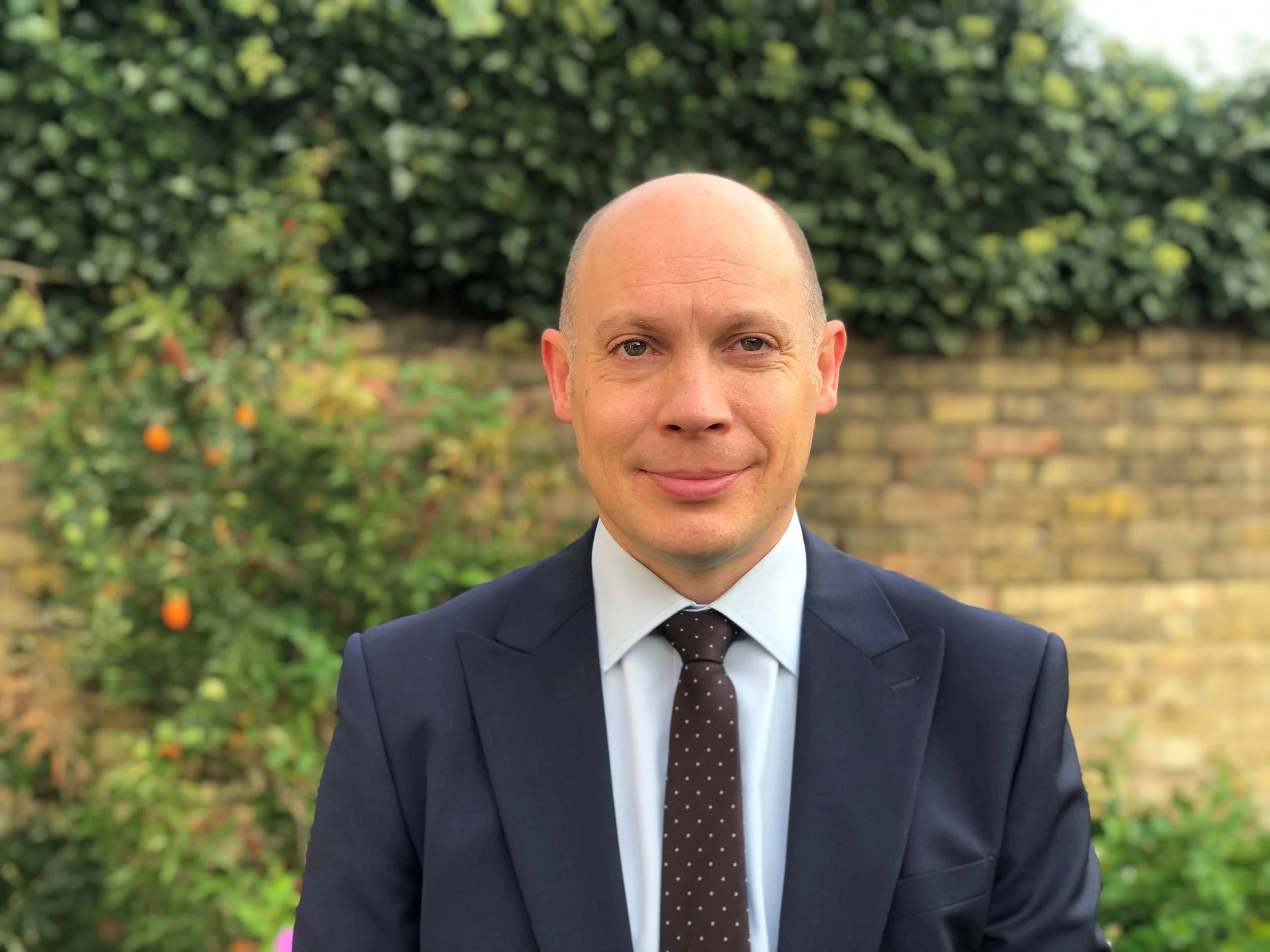 A man in a navy suit with a white shirt and polka dot tie is standing outdoors in front of a brick wall and green foliage.