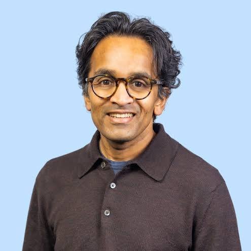Portrait of a man with glasses, dark hair, wearing a dark polo shirt against a light blue background.