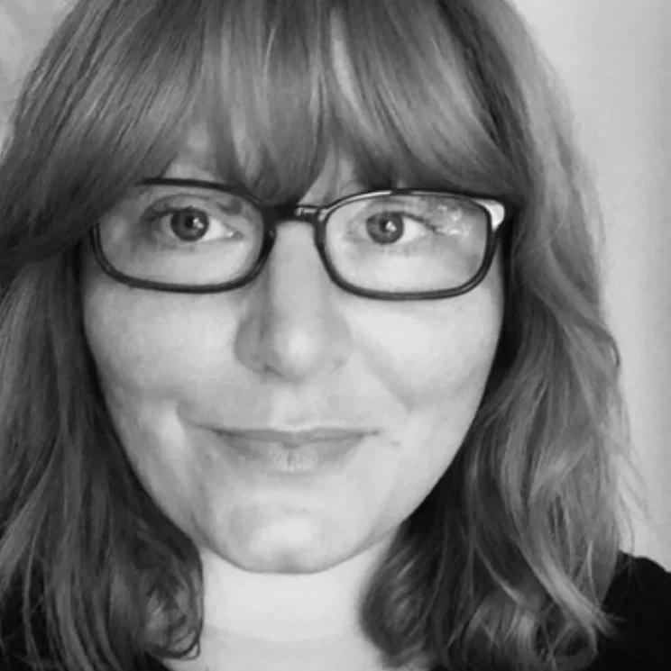 Black and white photo of a woman with shoulder-length hair, wearing glasses, smiling, with soft lighting and a plain background.