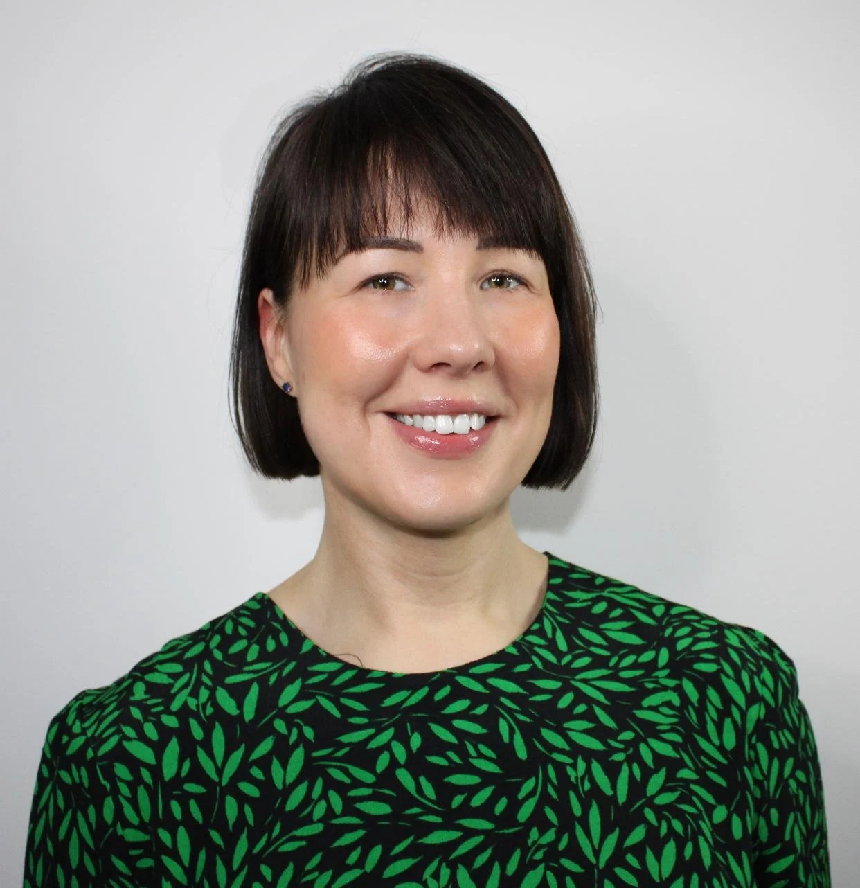 Headshot of a woman with short dark brown hair, wearing a green and black patterned top, smiling against a plain gray background.