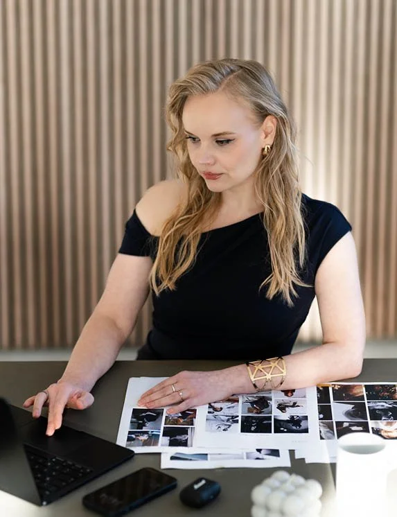 Eine blonde Frau sitzt in einem blauen Kleid am Schreibtisch und arbeitet am Laptop. Auf dem Schreibtisch befinden sich Dokumente, eine Kaffeetasse, ein Handy, Kopfhörer und eine Kerze. Im Hintergrund sitzt man eine Wand mit Akkustikpanelen aus Holz.