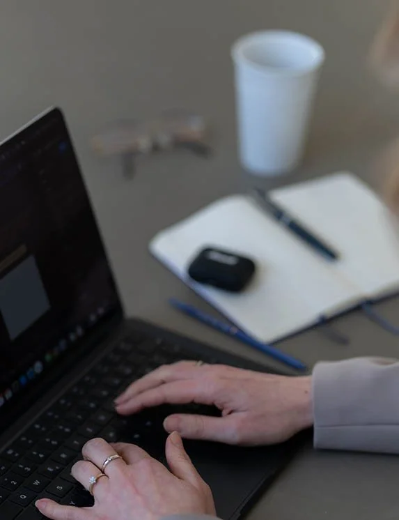 Wir Blicken auf einen Schreibtisch auf dem ein dunkler Laptop steht. Zwei weibliche Hände tippen auf einer Laptop-Tastatur. Im Hintergrund befindet sich ein aufgeschlagenes Notizbuch, ein Stift, eine Brille, Kopfhörer und eine Kaffeetasse.
