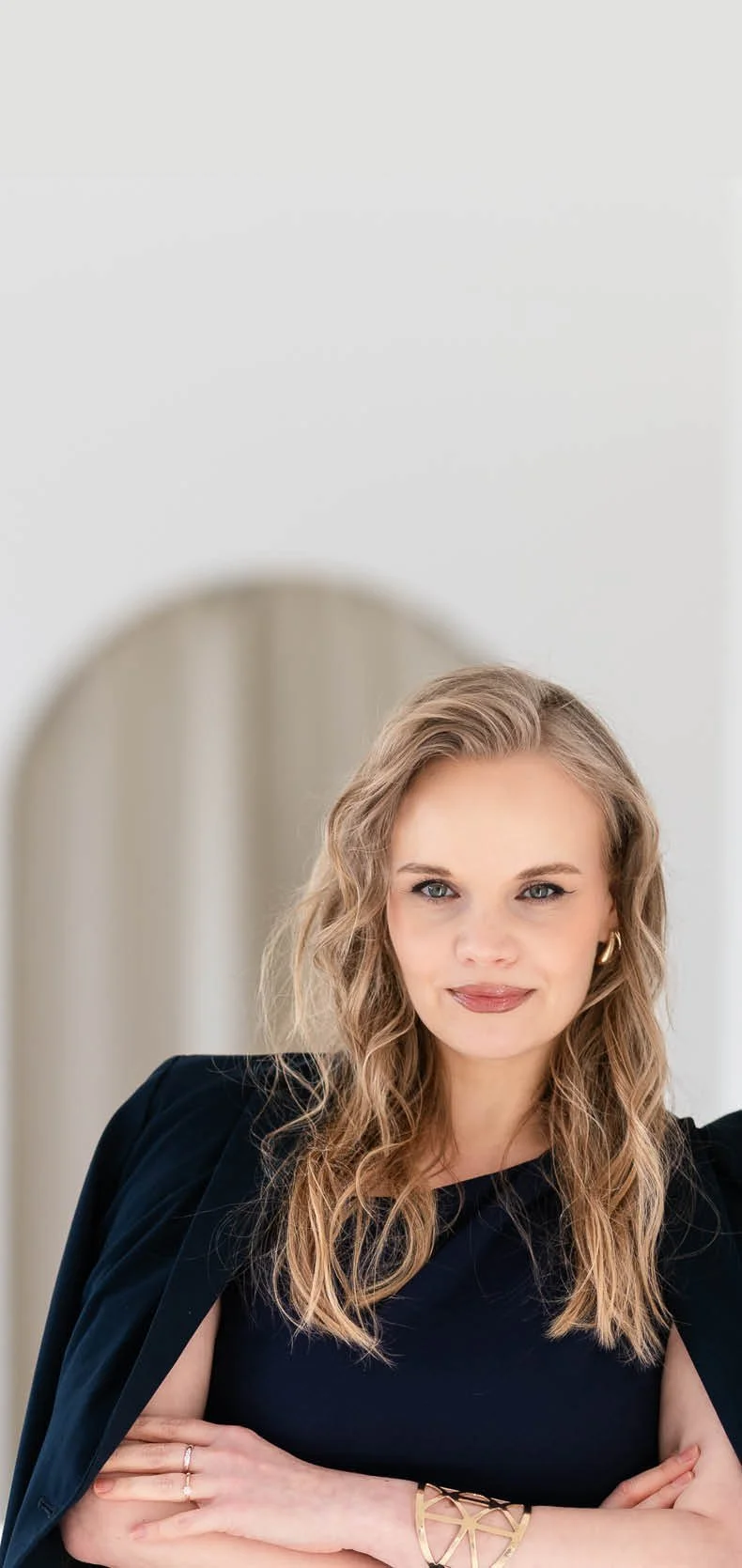 Eine Frau mit blondem gelockten Haar steht in einem hellen Raum an eine Wand gelehnt. Sie hat ein dunkel blaues Kleid an und über ihren Schultern liegt ein ebenfalls dunkel blauer Blazer. Sie hat die Arme vor der Brust gekreuzt und lächelt.
