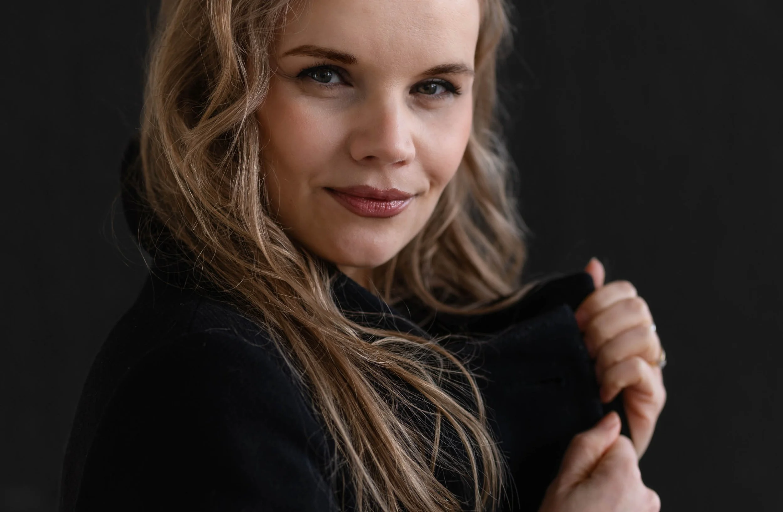 Eine blonde Frau mit blauen Augen schaut lächelnd in die Kamera und hält den Kragen eines schwarzen Blazers hoch. Der Hintergrund ist dunkel und unscharf.