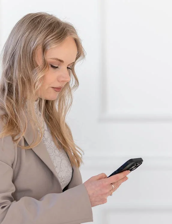 Eine blonde Frau in einem beigen Anzug mit einem weißen Spitzenoberteil steht und schaut auf ihr Smartphone, das sie in ihrer Hand hält. Im Hintergrund kann man einen hellen Raum erahnen.