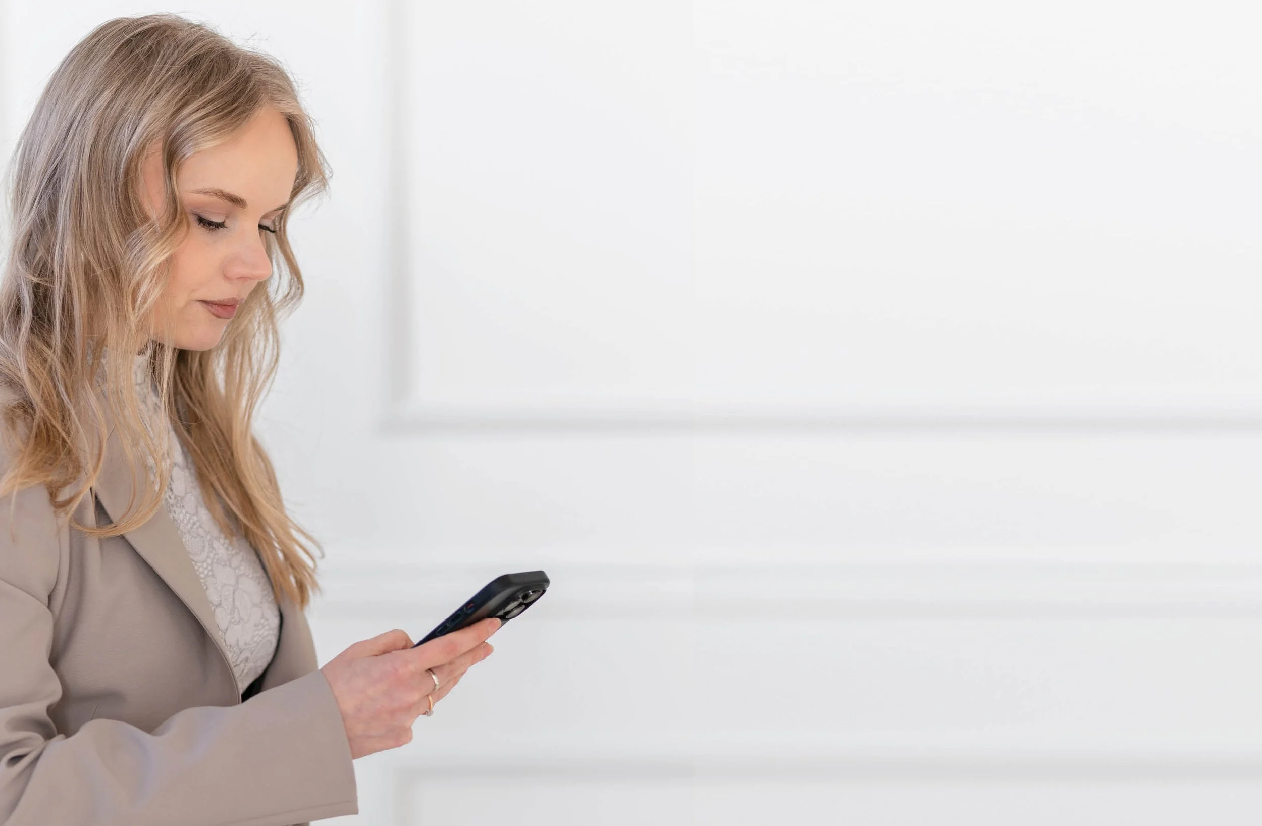 Eine Frau mit langen blonden lockigen Haaren steht in einem hellen Raum. Sie trägt einen beigen Anzug und ein weißes Oberteil. Ihr Blick ist nach unten auf ihr Smartphone gerichtet, welches sie in ihrer rechten Hand hält.
