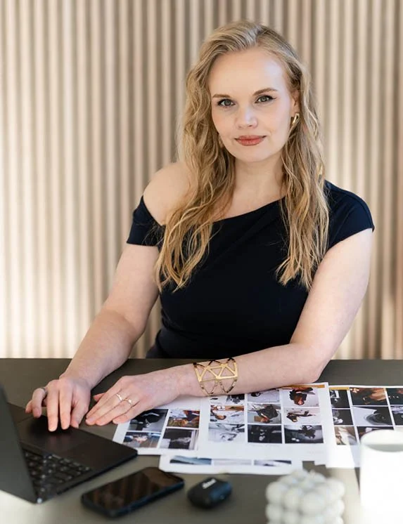 Eine blonde Frau in einem blauen Kleid mit goldenem Armreifen sitzt am Schreibtisch und ihre Finger liegen auf der Tastatur eines dunklen Laptops. Sie schaut lächelnd in die Kamera. Auf dem Schreibtisch liegen Dokumente, ein Smartphone, Kopfhörer.