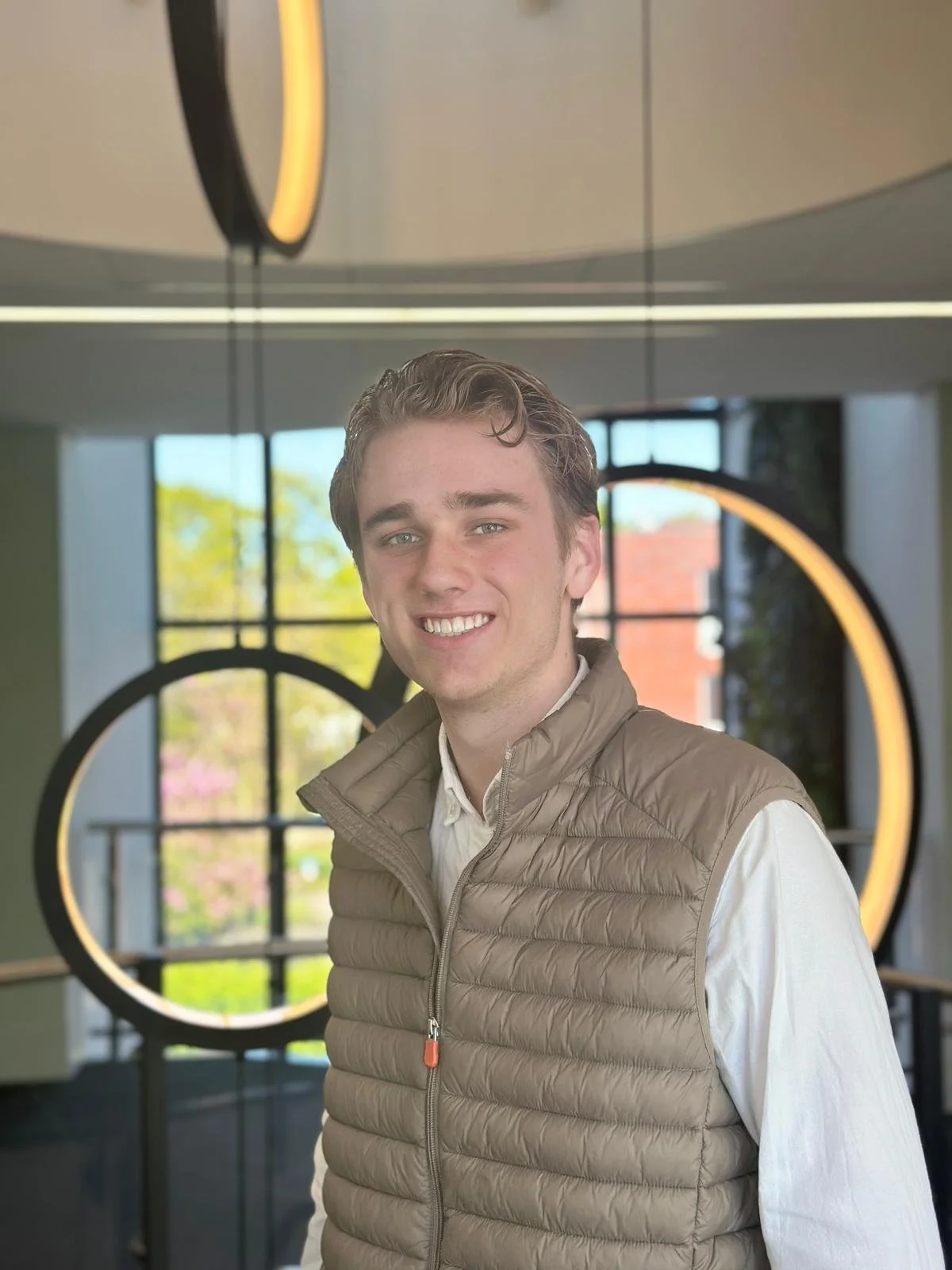 A young man with brown hair and a light complexion smiling, standing indoors with modern circular light fixtures and large windows showing a green outdoor scene in the background.