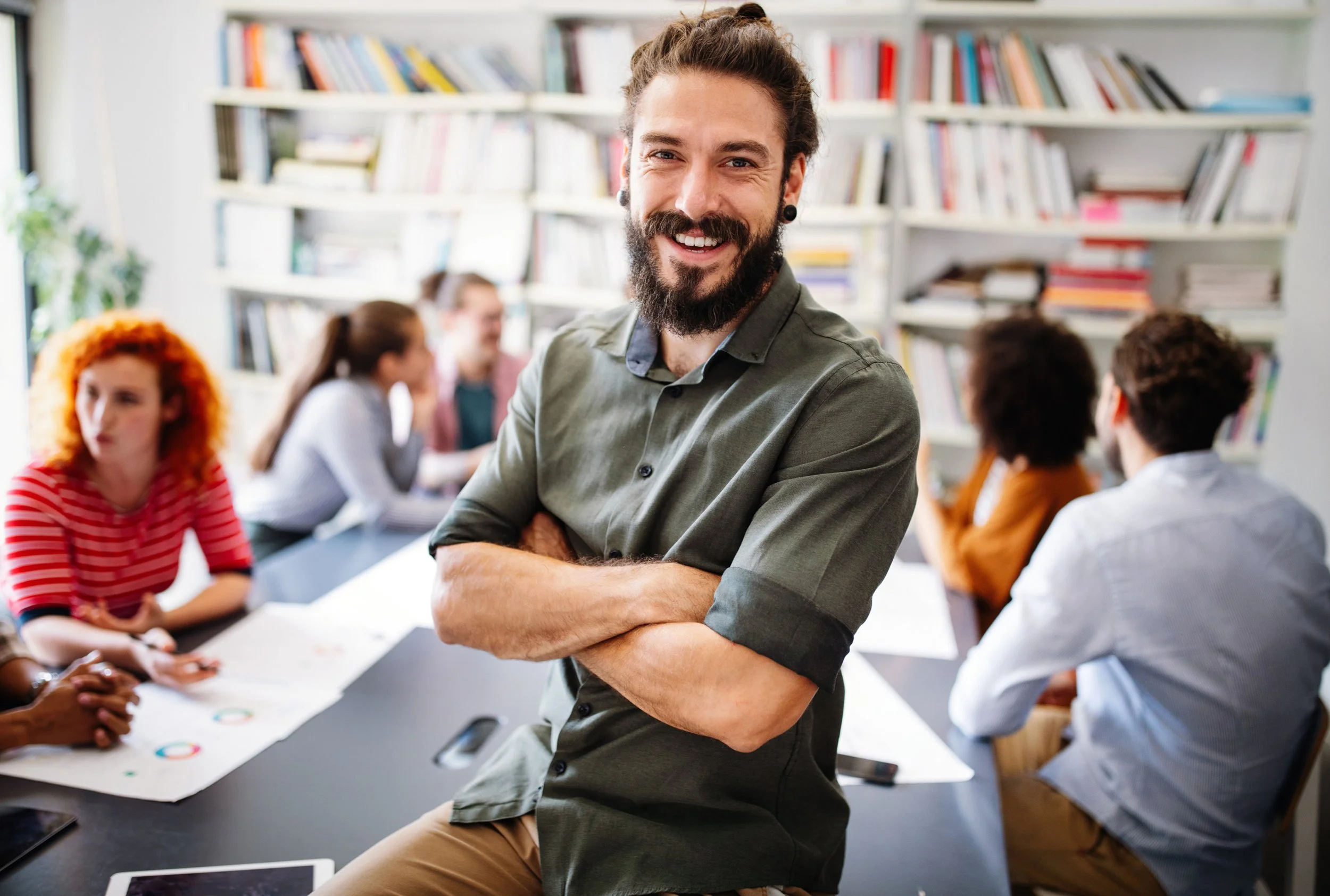 A smiling man with a beard and earrings stands with his arms crossed in a library or classroom, with a group of people sitting at a table in the background.