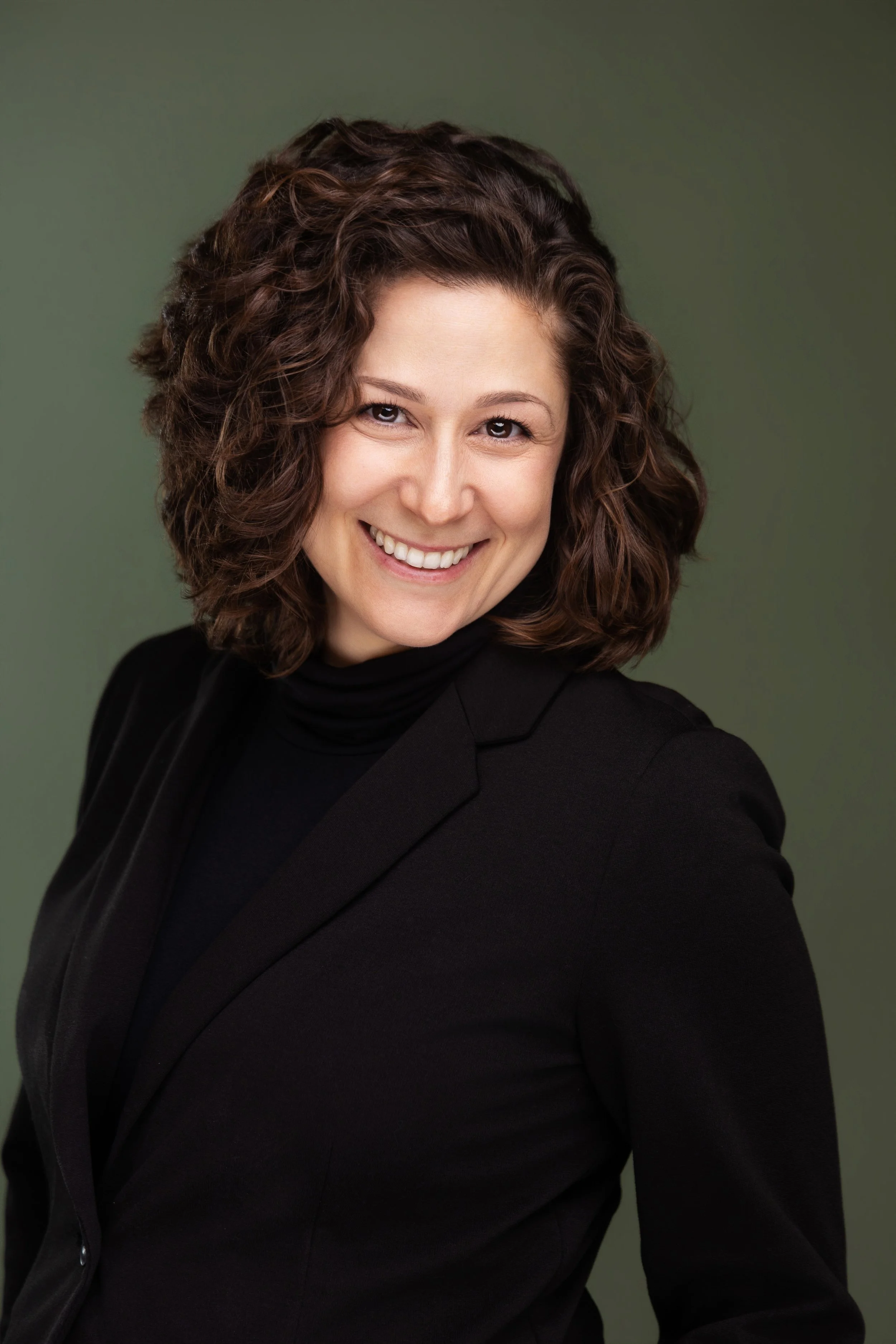 Headshot of Dr. Melissa Greene with curly brown hair, smiling, wearing a black blazer and turtleneck, against a green background.