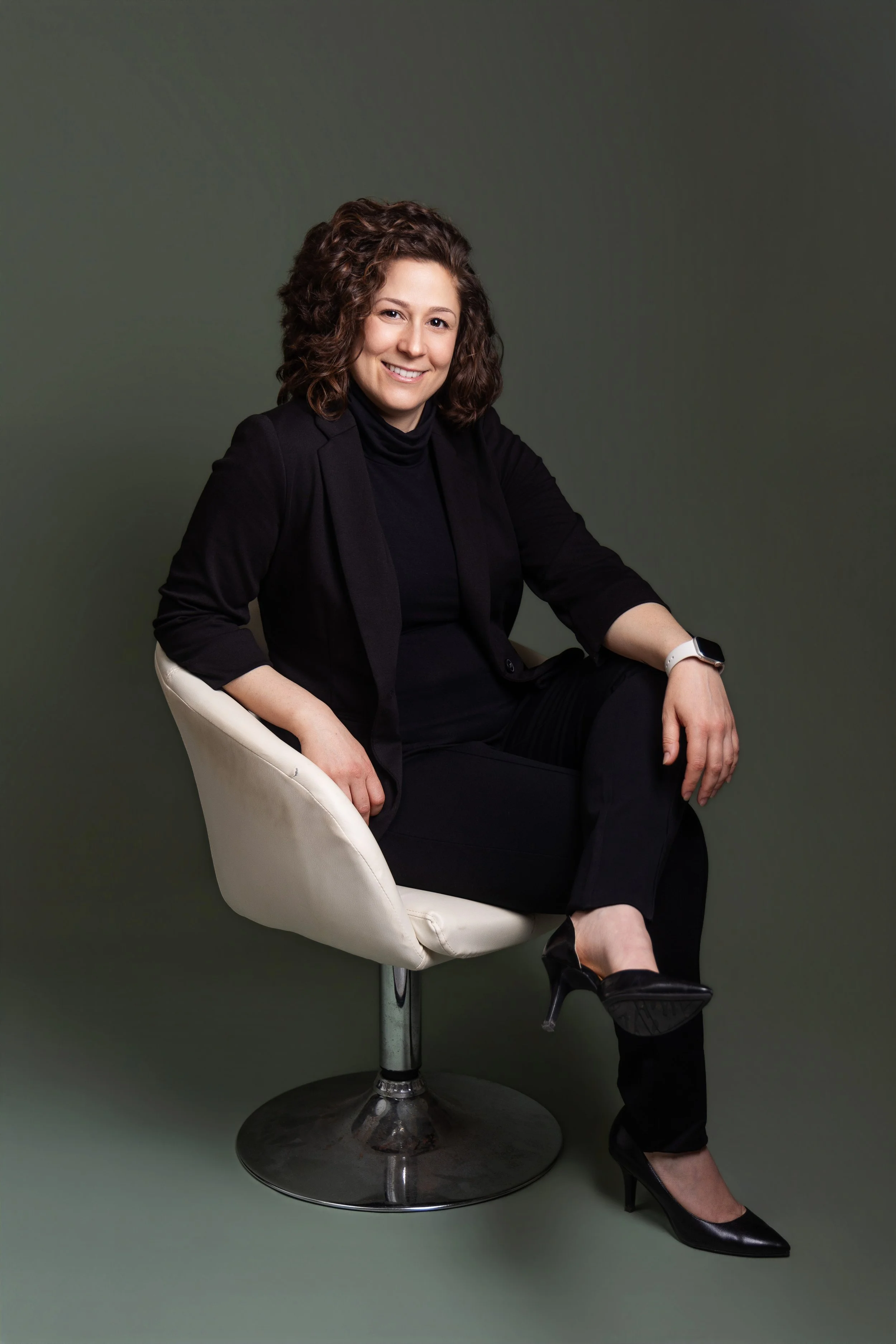 Dr. Melissa Greene smiling, wearing a black blazer, black turtleneck, black pants, black high heels, and a white watch, sitting in a white modern chair against a dark green background.