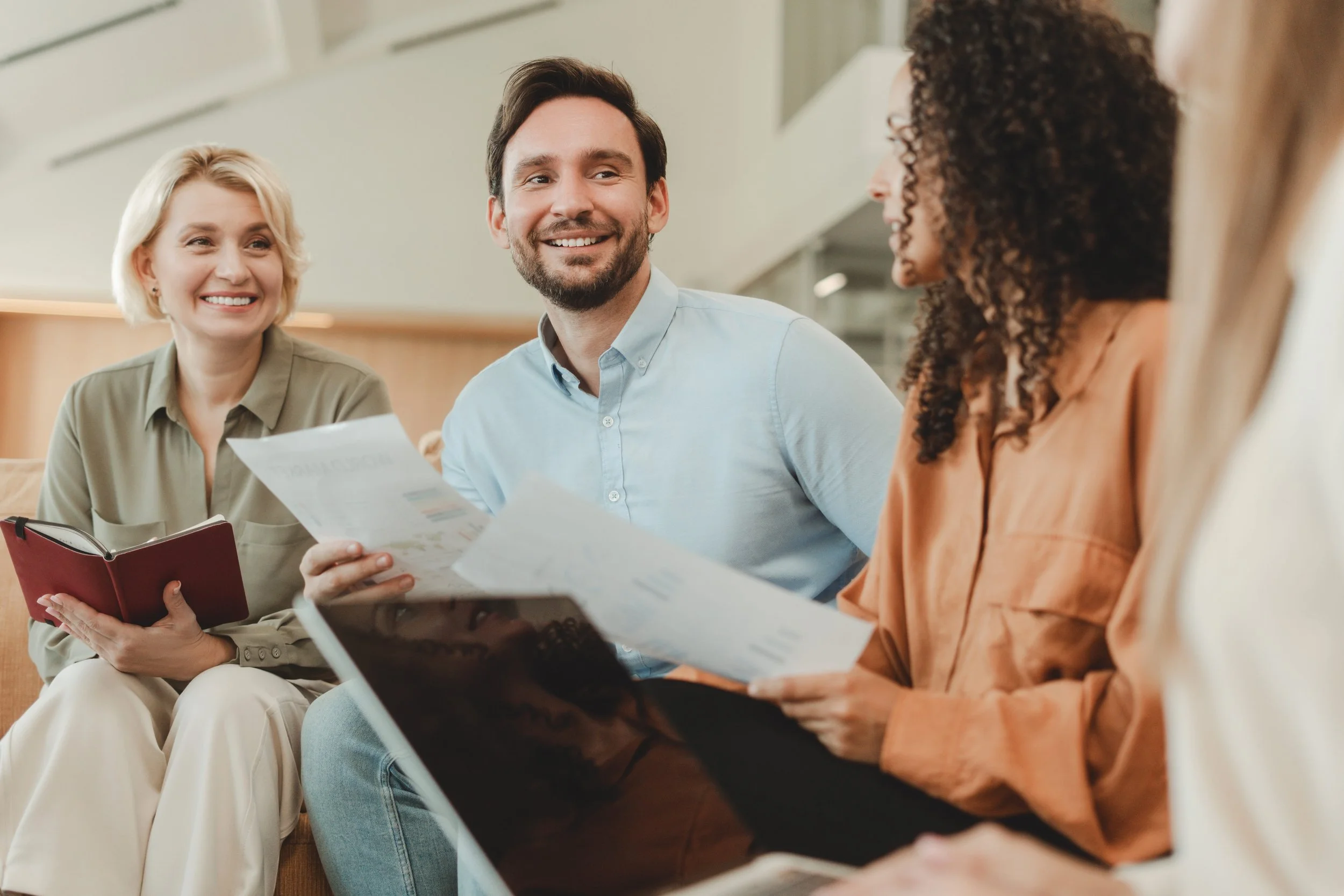 A diverse group of three people sitting together in a professional setting, engaged in a discussion. The woman on the left has short blonde hair and is holding a red notebook, smiling. The man in the middle has dark hair and a beard, and is smiling while holding some papers. The woman on the right has curly hair, wearing an orange top, and is holding a contract or document. The background shows an indoor space with neutral decor.