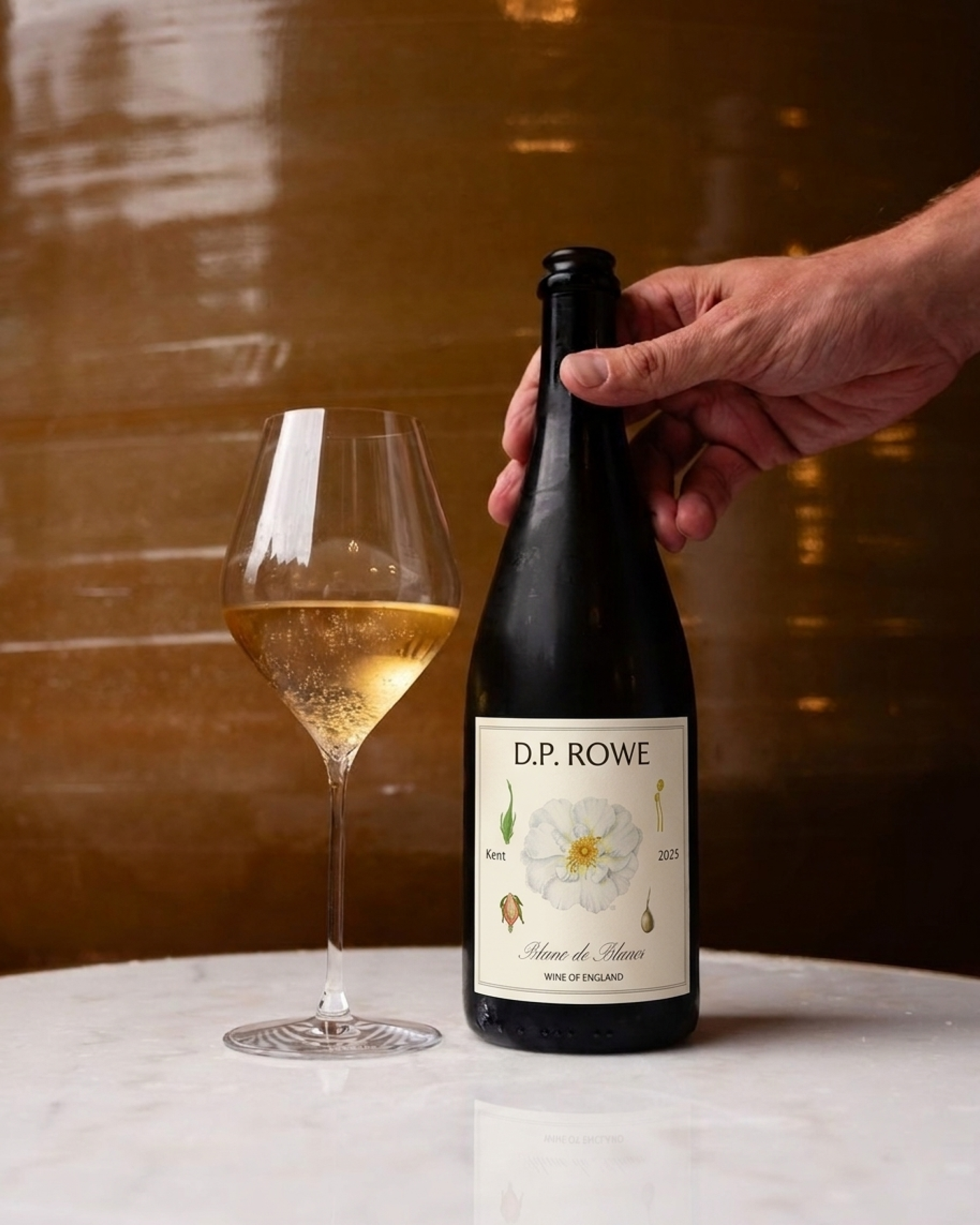 A hand opening a black bottle of white wine labeled 'D.P. ROWE' with a glass of white wine next to it on a reflective white surface, against a warm blurred background.