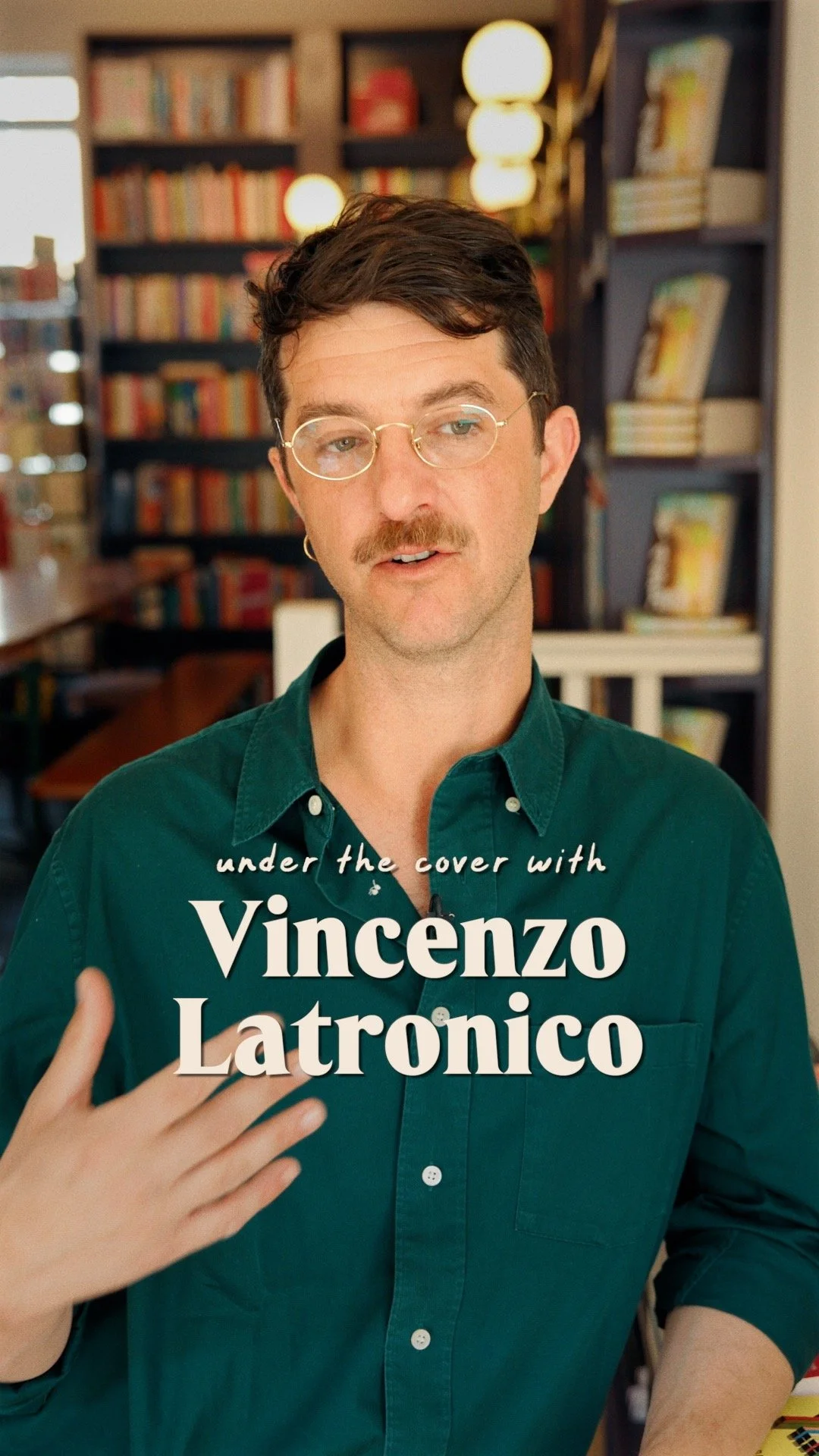 A man wearing glasses and a green button-up shirt, gesturing with his right hand, standing in front of a bookshelf filled with books and warm lighting.
