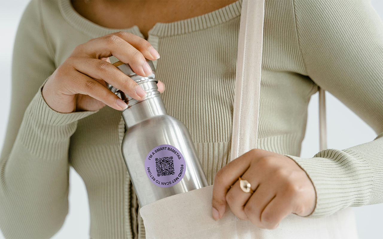 Person holding a stainless steel water bottle with a QR code sticker on it, wearing a beige ribbed long sleeve shirt and a gold ring.