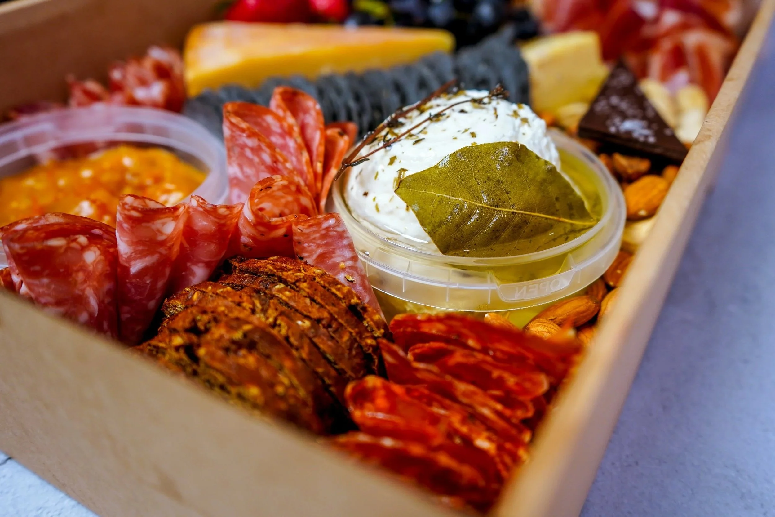 A box of assorted cured meats, cheeses, crackers, a container of mustard, a dollop of whipped cream with herbs, and a bay leaf, arranged for a snack.