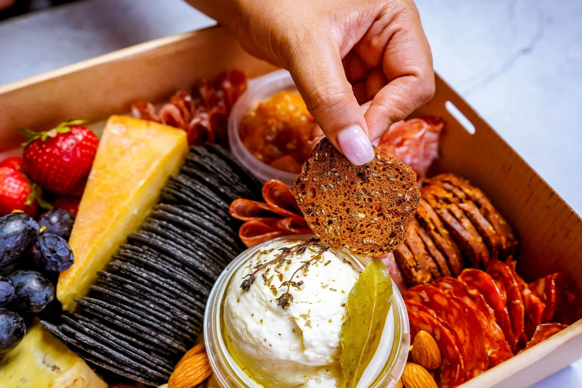 Hand selecting a cracker from a beautifully presented grazing box filled with artisan cheeses, cured meats, fresh fruit and gourmet produce.