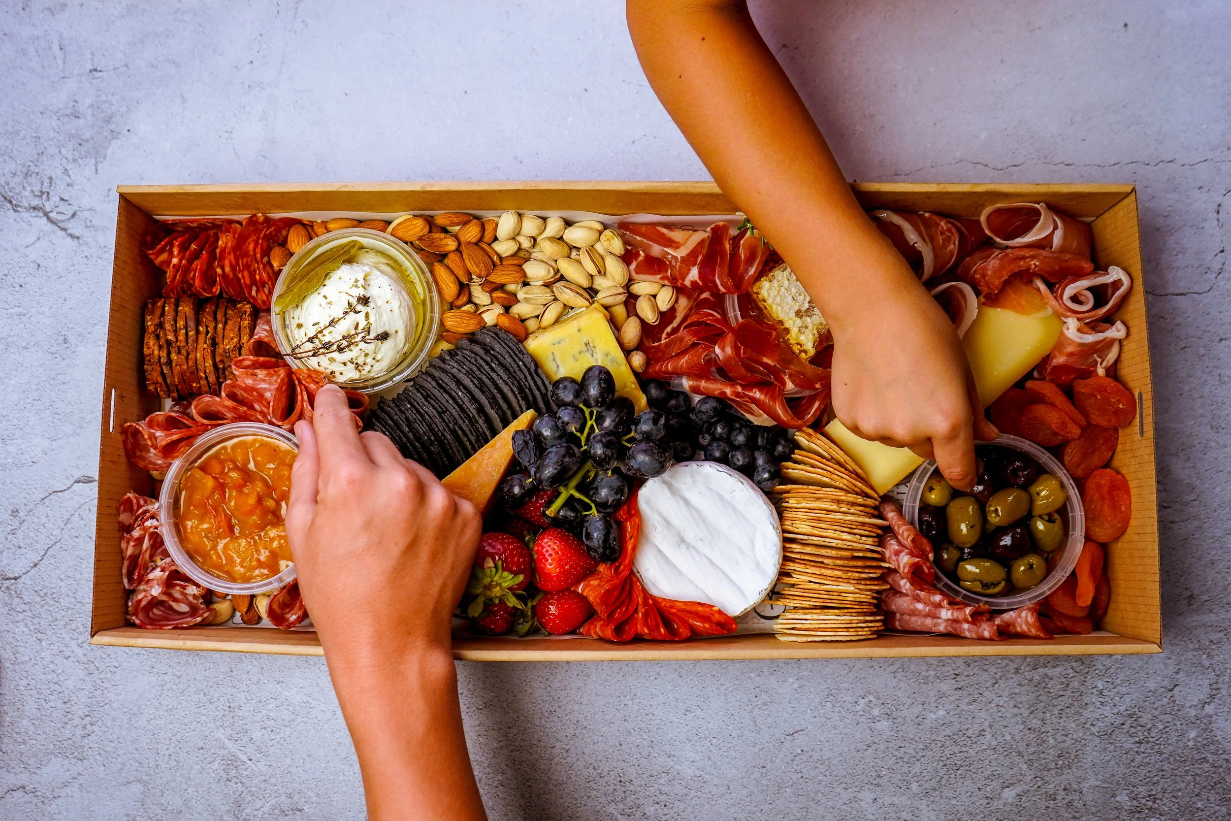 A cheese and charcuterie board with various cheeses, cured meats, olives, grapes, strawberries, almonds, pistachios, and crackers, presented in a rectangular wooden box.