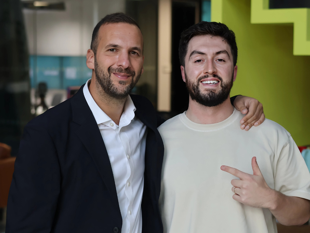 Two men posing together indoors, one in a dark suit jacket and white shirt, the other in a light t-shirt, with the man in the t-shirt pointing at himself and the other man smiling. Zack Polanski and Jimmy the Giant.