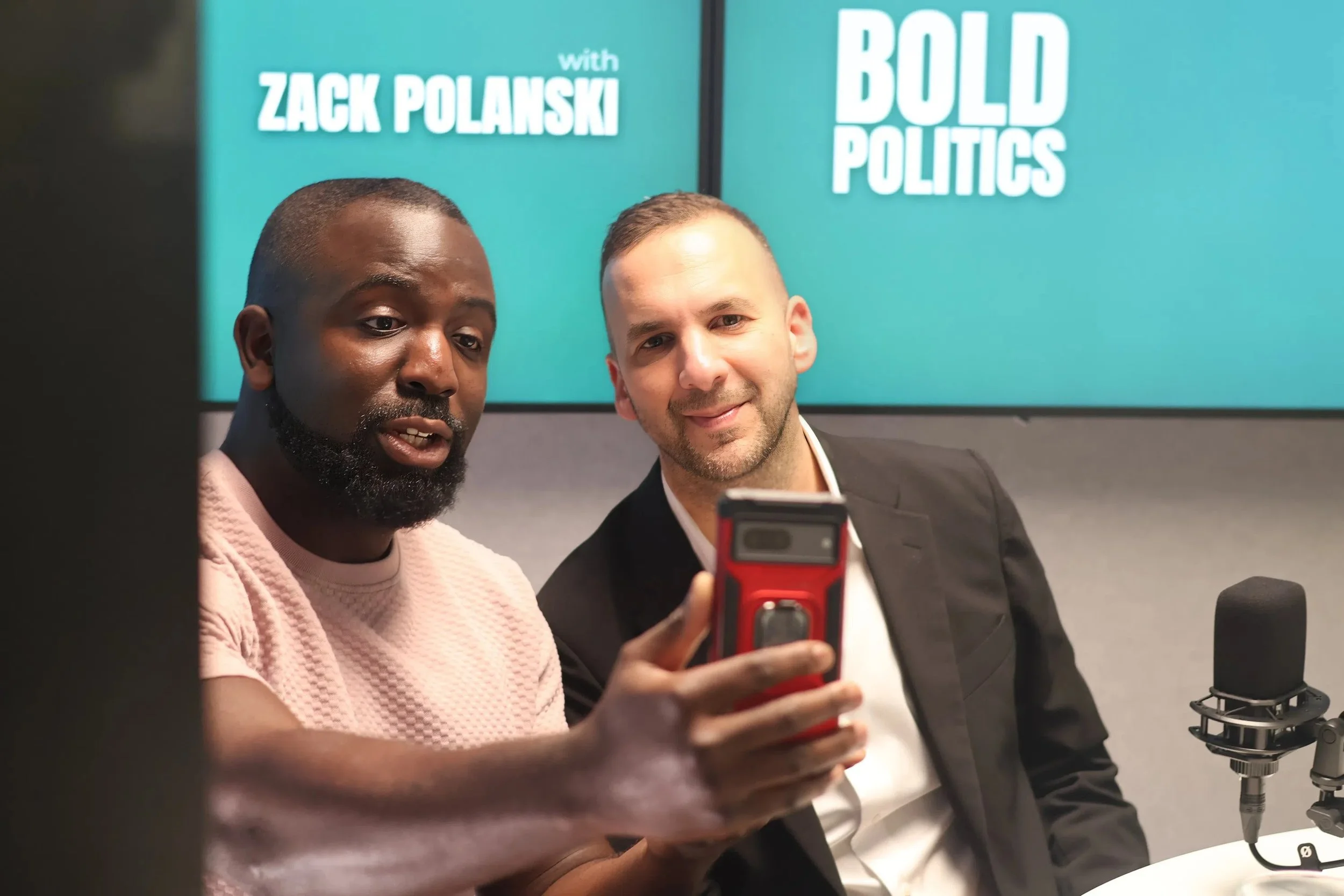 Two men in a recording studio, one wearing a light pink shirt and the other in a black blazer, looking at a smartphone together. Behind them are two screens displaying 'ZACK POLANSKI' and 'BOLD POLITICS'.