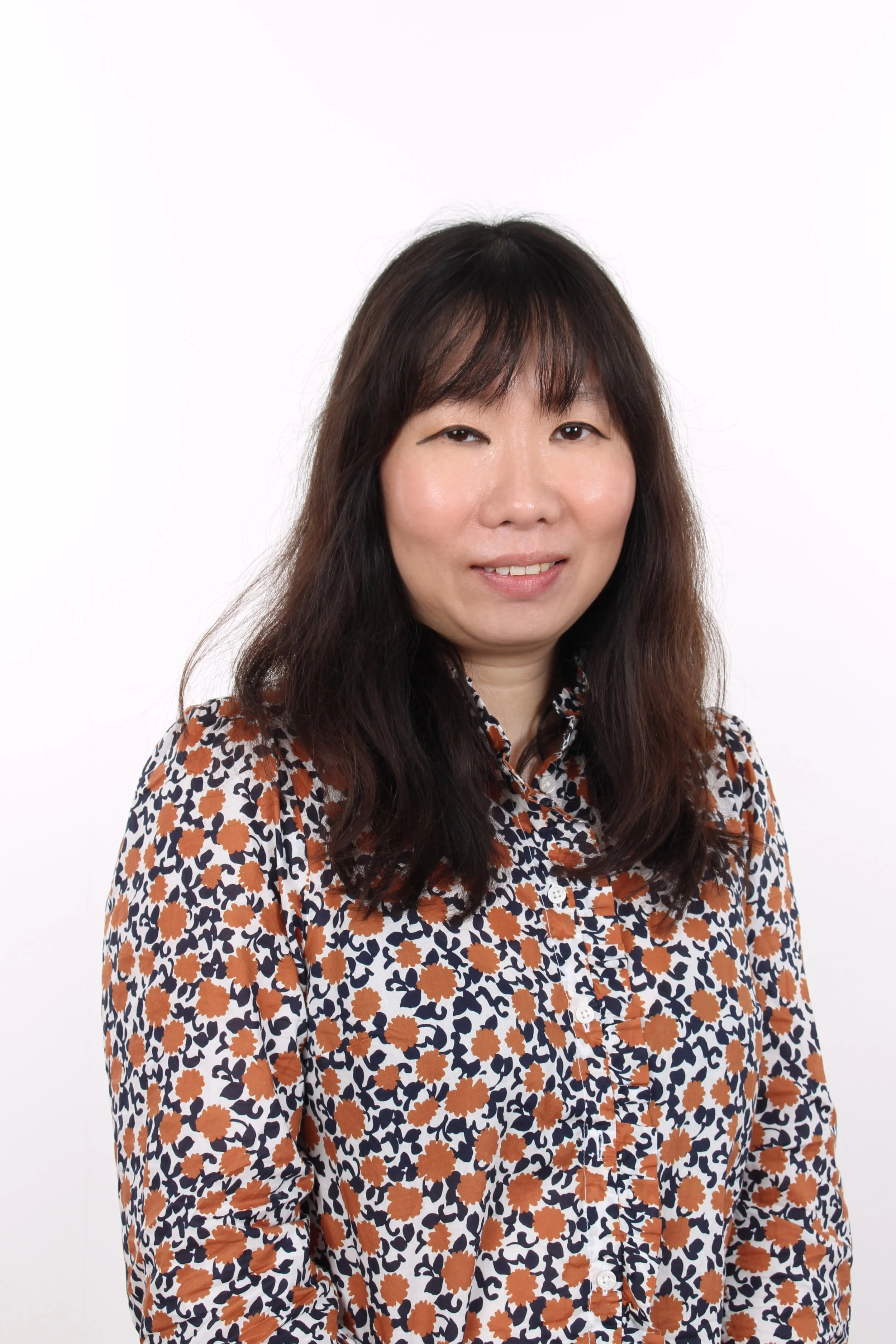 A woman with dark brown hair and a patterned shirt standing against a white background.