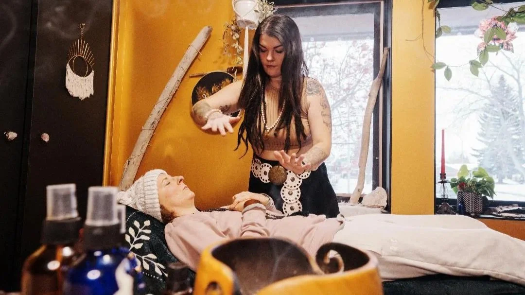 A woman with long dark hair, tattoos, and dressed in earthy tones stands next to a bed, gesturing with her hands, while a person lies on the bed covered with a pink blanket, wearing a knitted hat, and looking up at her. The room has yellow walls, a large window with snow outside, and various decorations including a moon wall hanging.