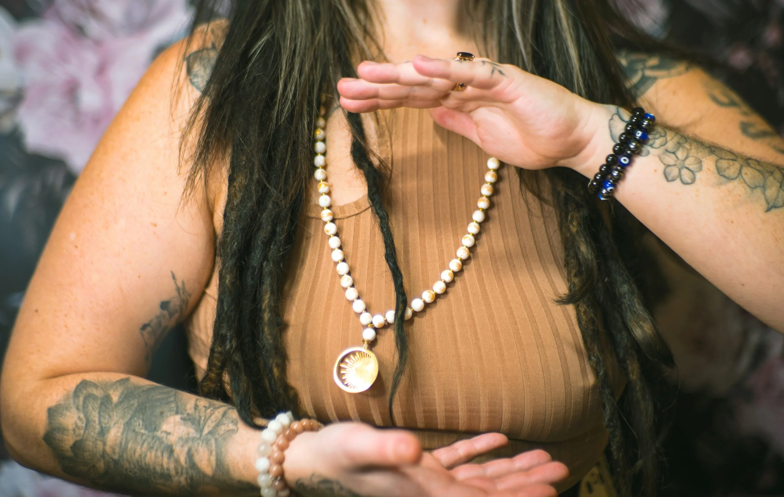 A woman with tattoos on her arms and long dreadlocks is wearing multiple necklaces and bracelets, holding her hand near her face in a gesture of invocation or blessing.