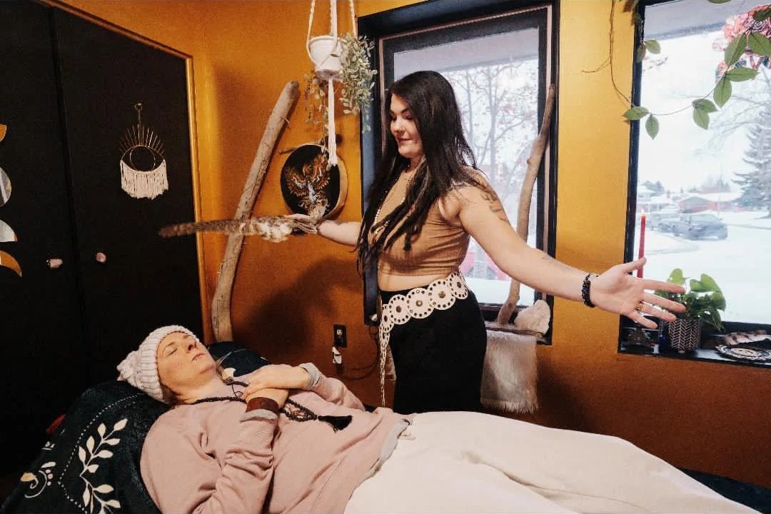 A person lying in bed with a white hat and pink sweater, appearing to be sleeping or resting, while another person stands nearby with arms outstretched and a bird on their hand, in a cozy room with a window showing snow outside.