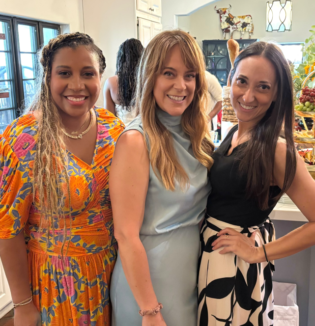 Three women smiling and standing together indoors, with a kitchen background and fruits on a counter.