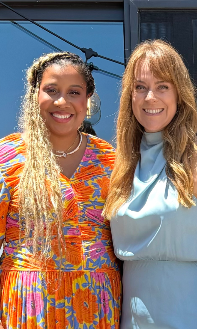 Two women smiling outdoors in front of a modern glass building, with a bright sunny atmosphere. The woman on the left has long, blonde, curly hair styled in twists and is wearing a colorful, floral patterned dress in orange, pink, and blue. The woman on the right has long, straight, light brown hair and is dressed in a light blue sleeveless top. Both women appear happy and relaxed.