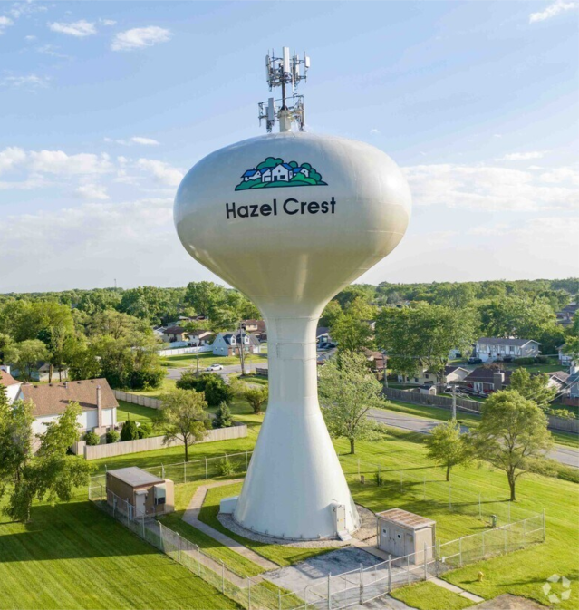 Water tower with Hazel Crest logo surrounded by residential neighborhood and green trees.