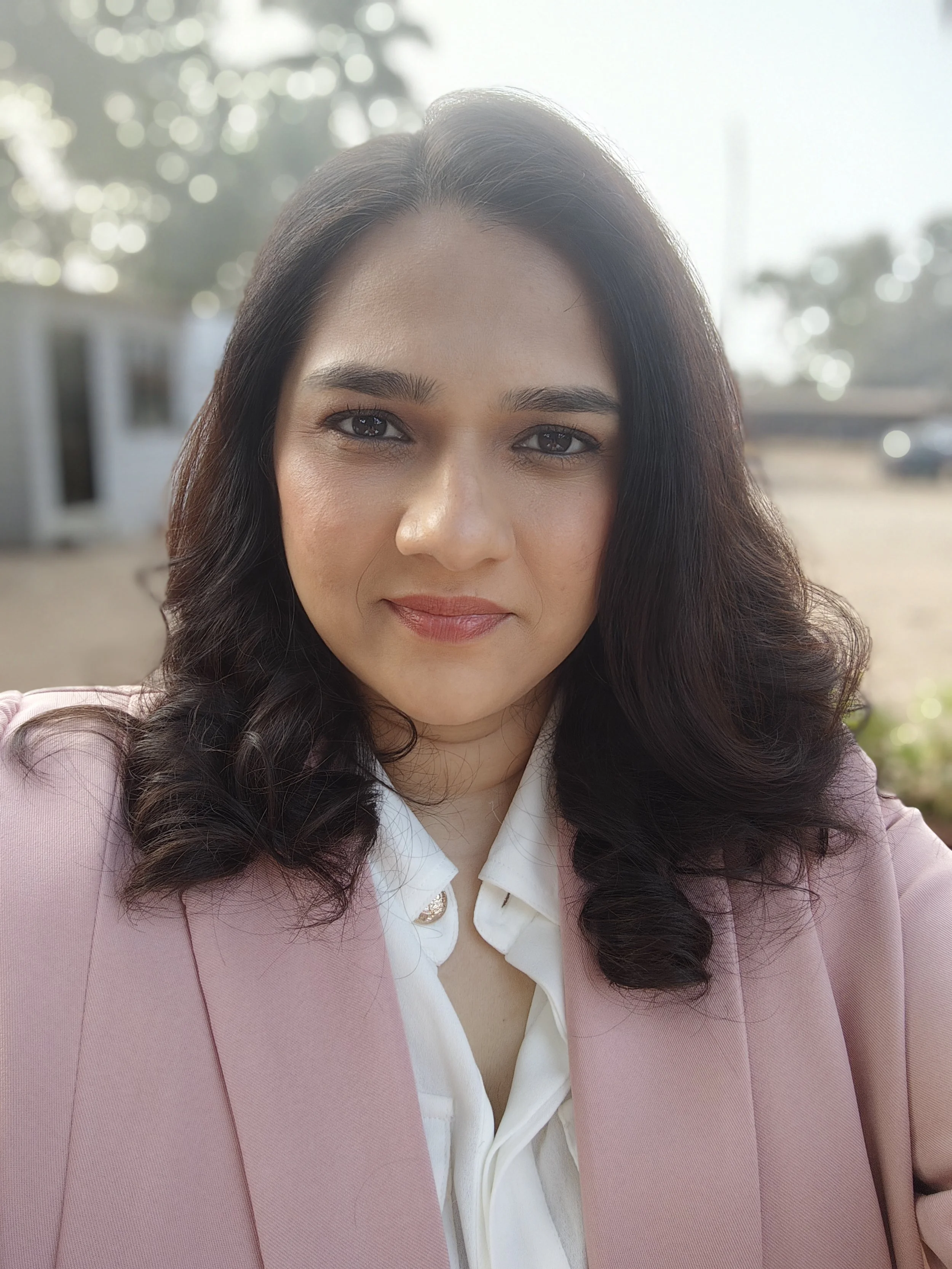 A woman with dark hair styled in loose curls, wearing a pink blazer over a white button-up shirt, taking a selfie outdoors in bright sunlight.