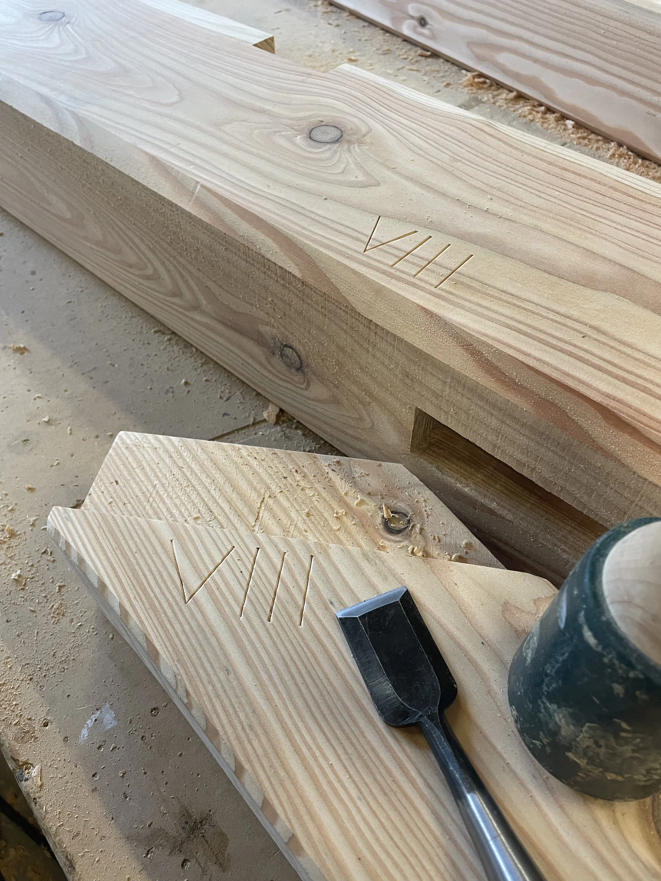 Two pieces of wood with the initials 'VHI' carved into them, a hammer, and a black marker on a workbench at a woodworking shop.