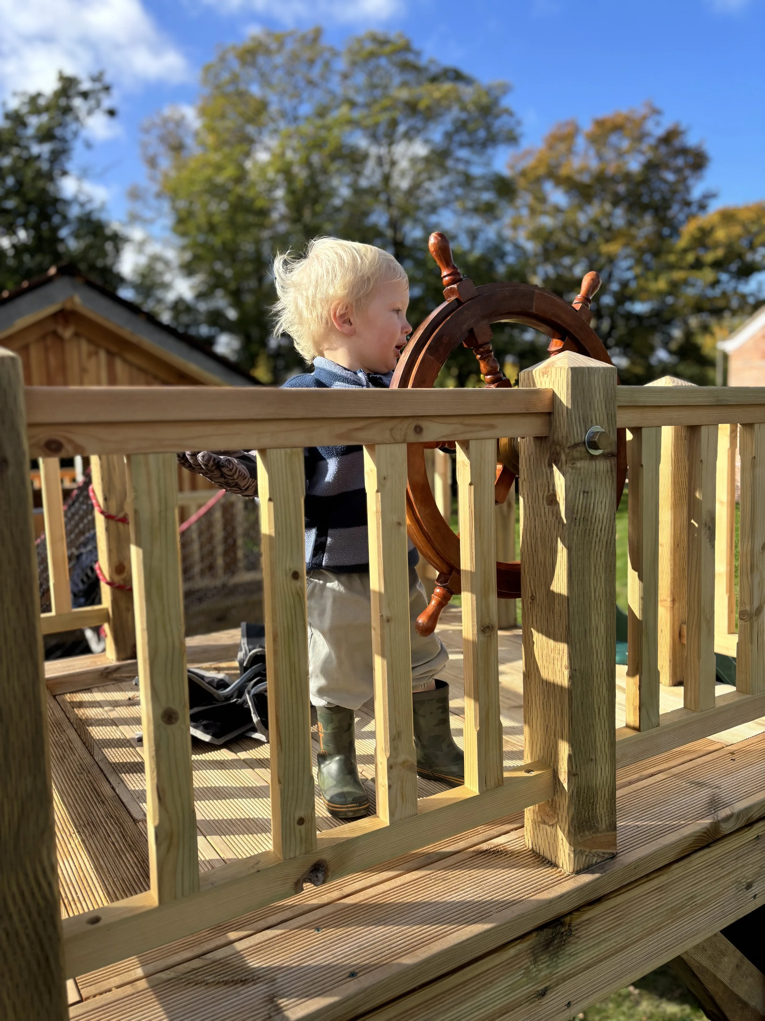 A young boy with blonde hair standing on a wooden deck, holding a ship's wheel. He is wearing rain boots, beige pants, a striped shirt, and a blue jacket. The deck is outdoors with trees and a house in the background, and the sky is partly cloudy with sunlight.