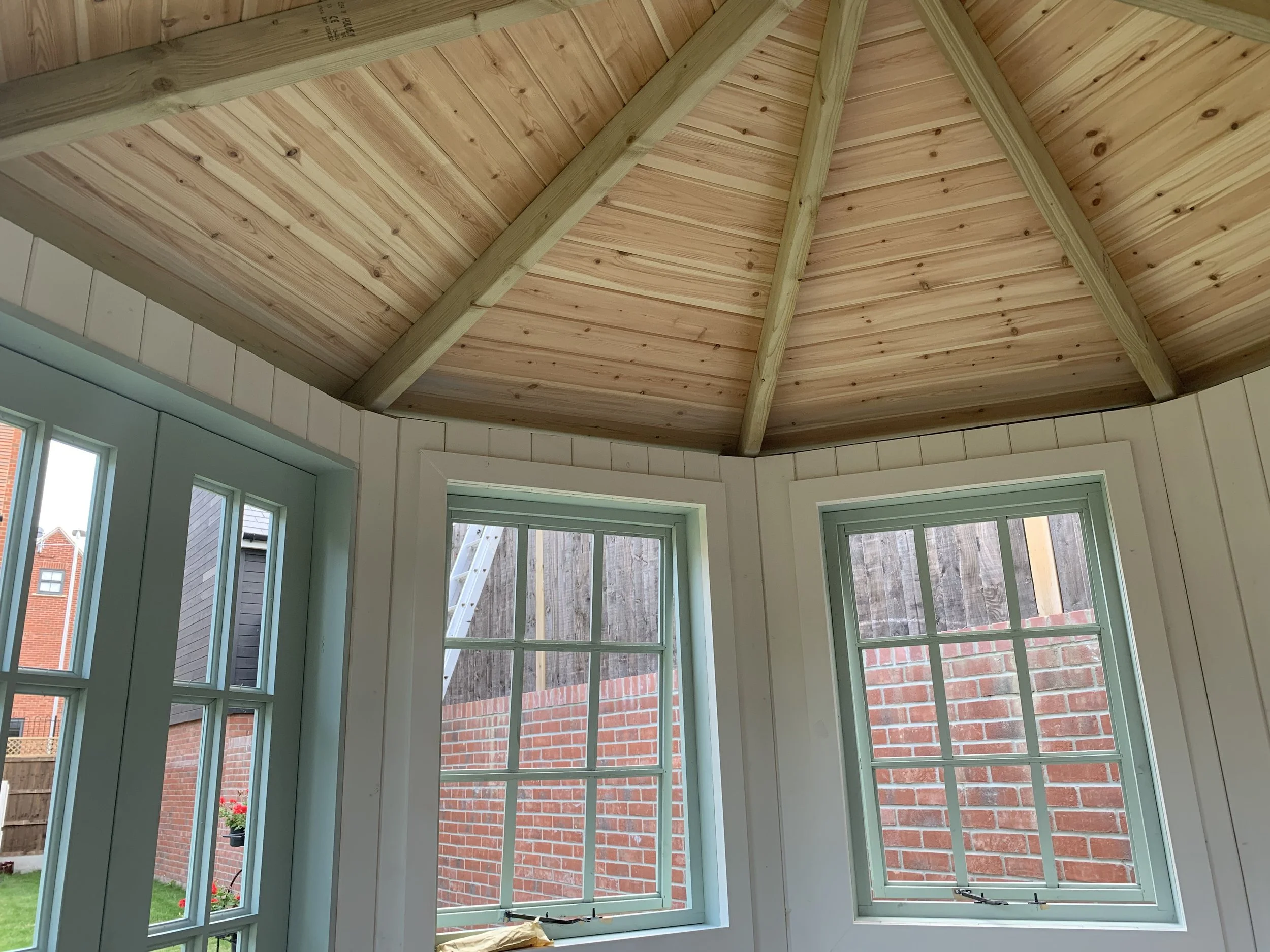 Interior view of a room with a wooden, vaulted ceiling and three green-framed windows facing a brick wall outside.