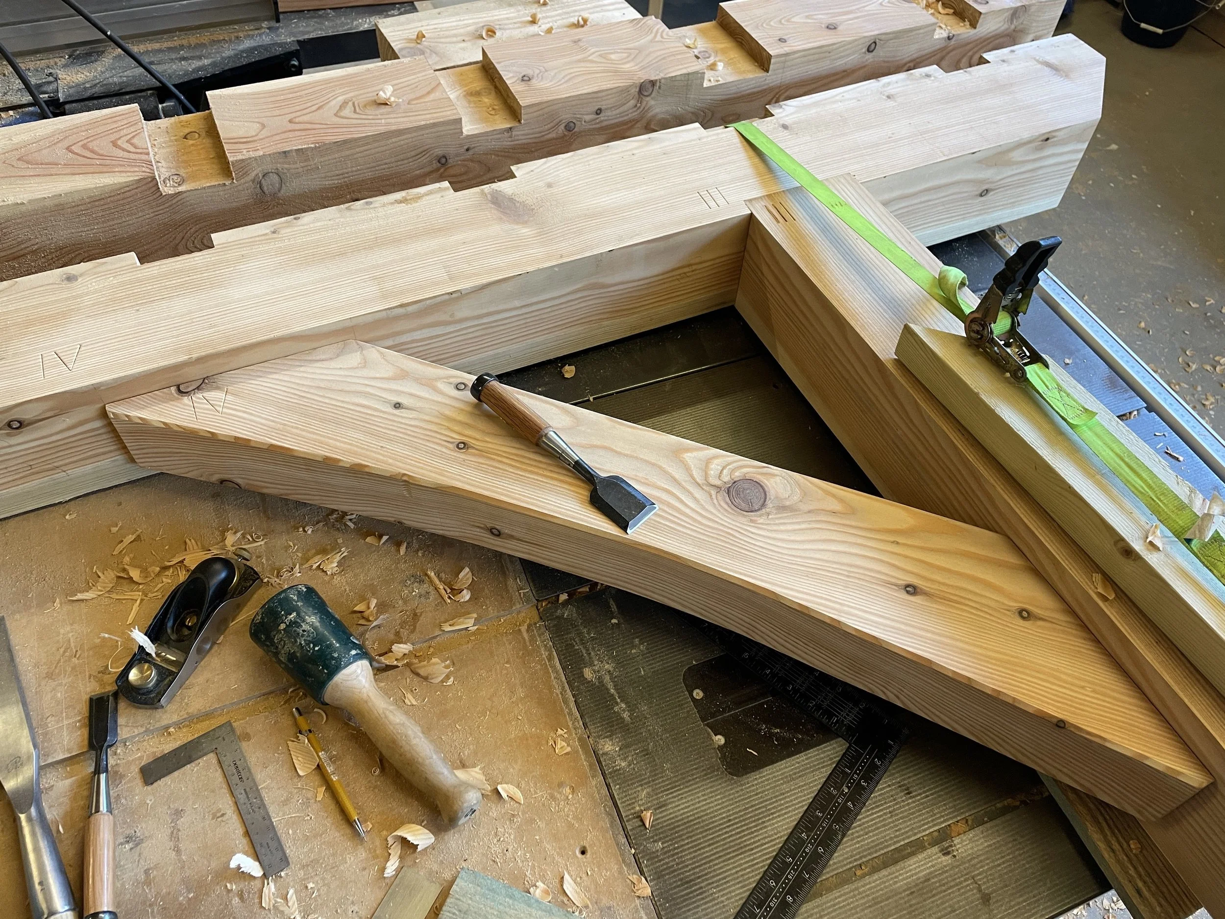 Woodworking workshop with a large wooden beam clamped in place for cutting or shaping. Tools such as a chisel, hammer, square, and clamps are visible on a workbench, along with wood shavings and protective tape.