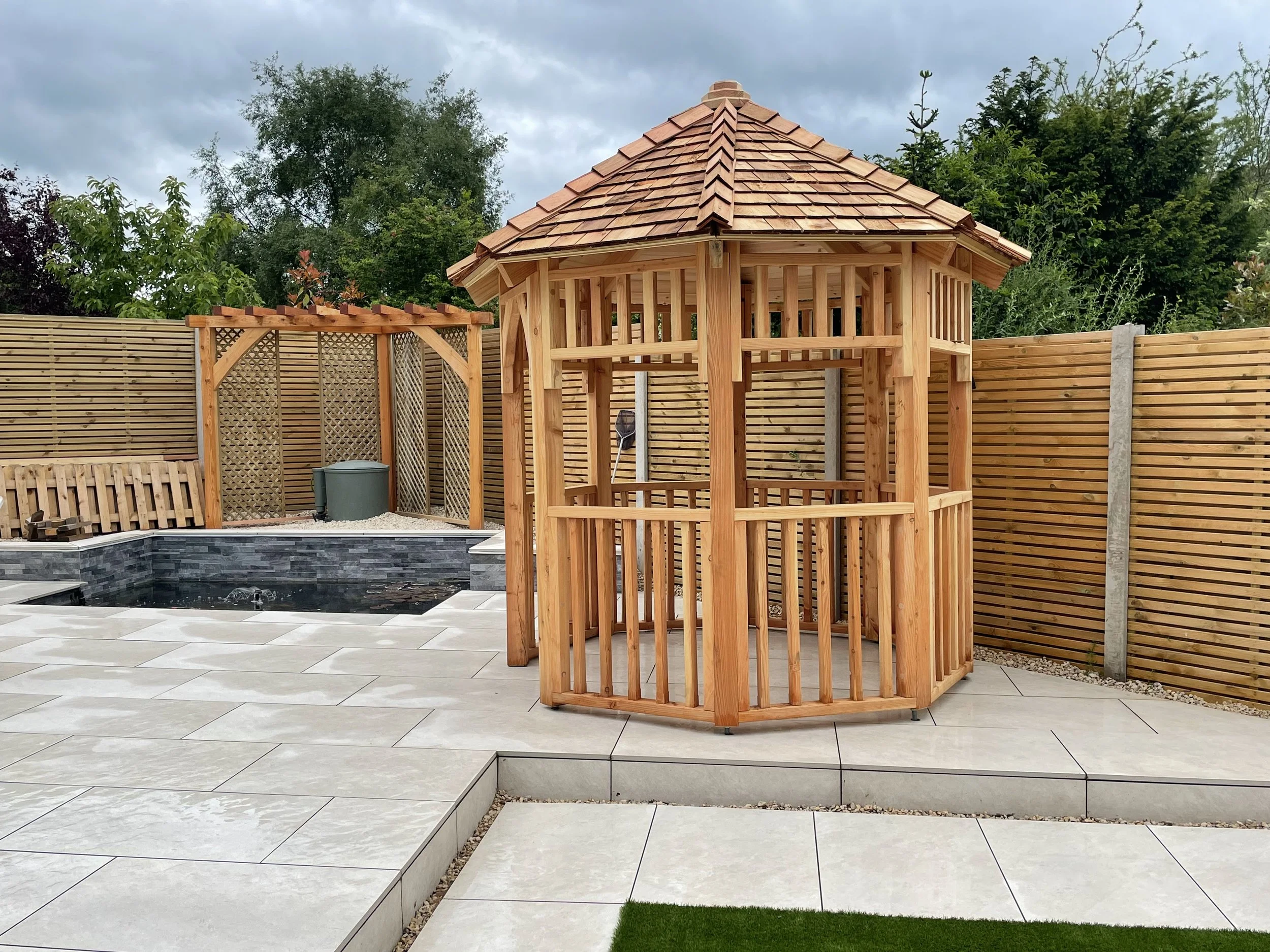 A wooden gazebo with a shingled roof in a backyard, surrounded by a wooden fence, with a small pool and a privacy screen in the background.