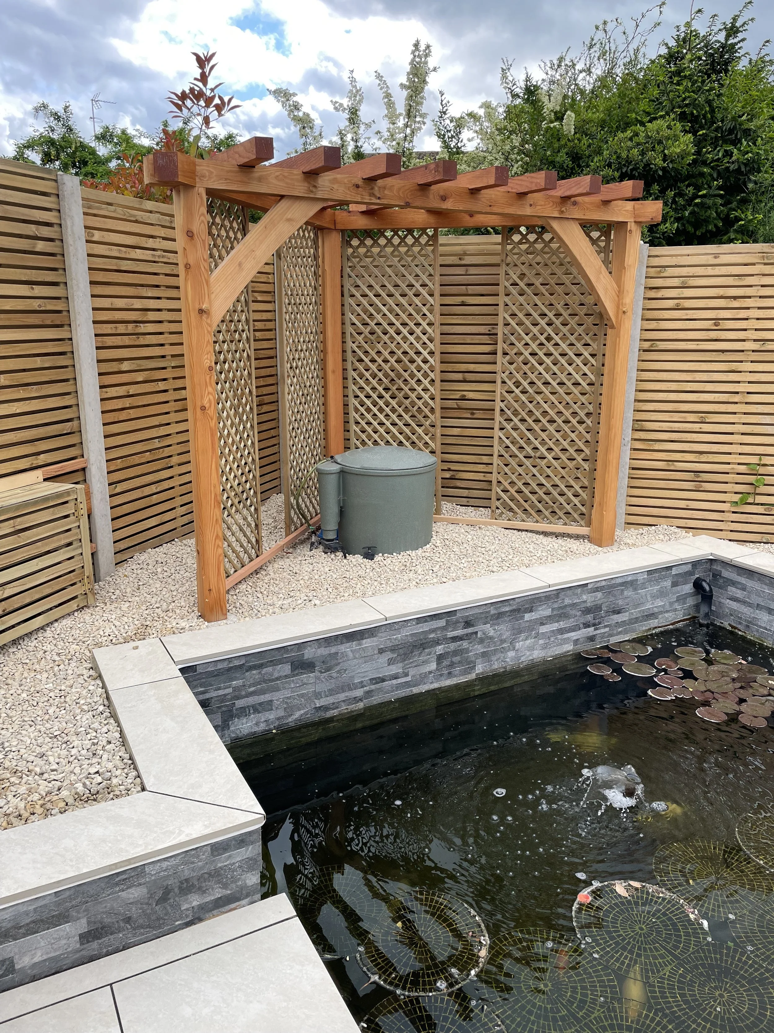 Backyard with a pond, water lilies, a wooden structure for an outdoor shower, and a gravel area with a green water filter.