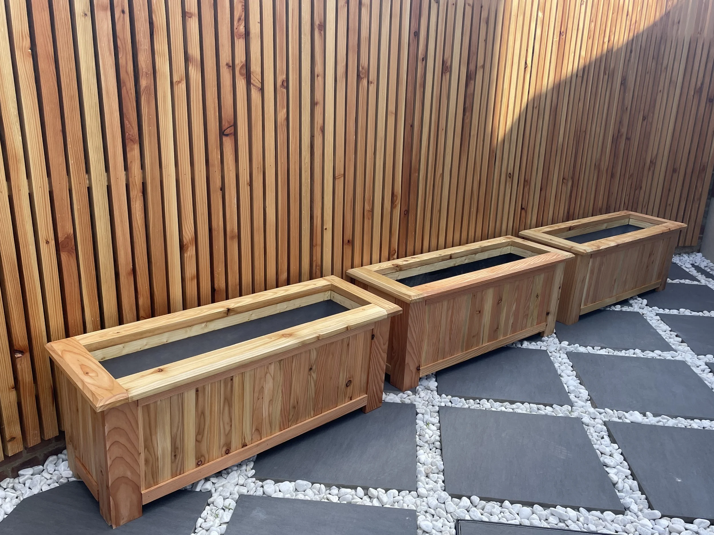Three rectangular wooden planters with black lining, placed on a stone-paved area against a wooden fence wall.