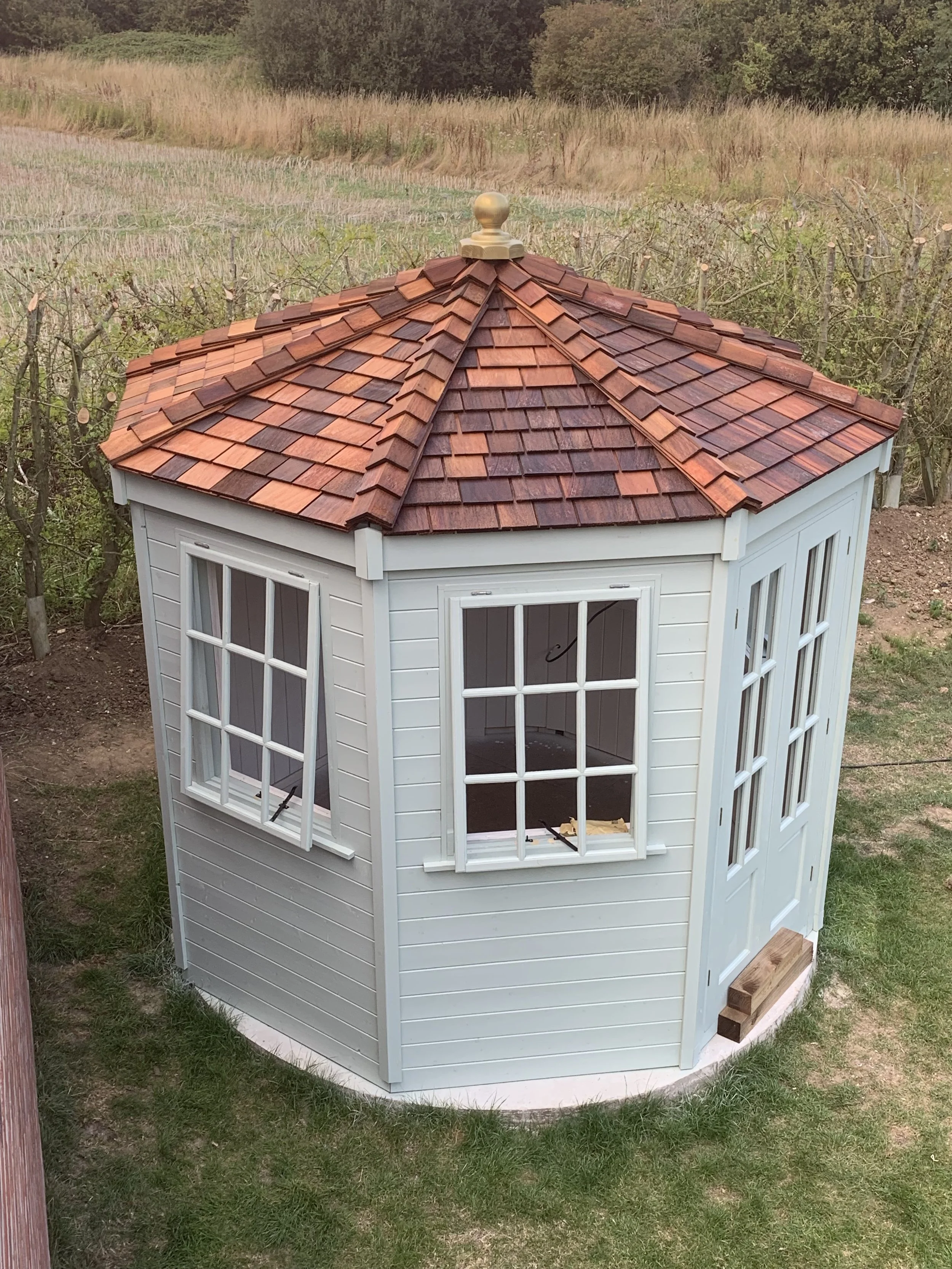 A small white wooden garden shed with a red shingled roof and multiple windows, situated on a grassy area near a vineyard or garden.