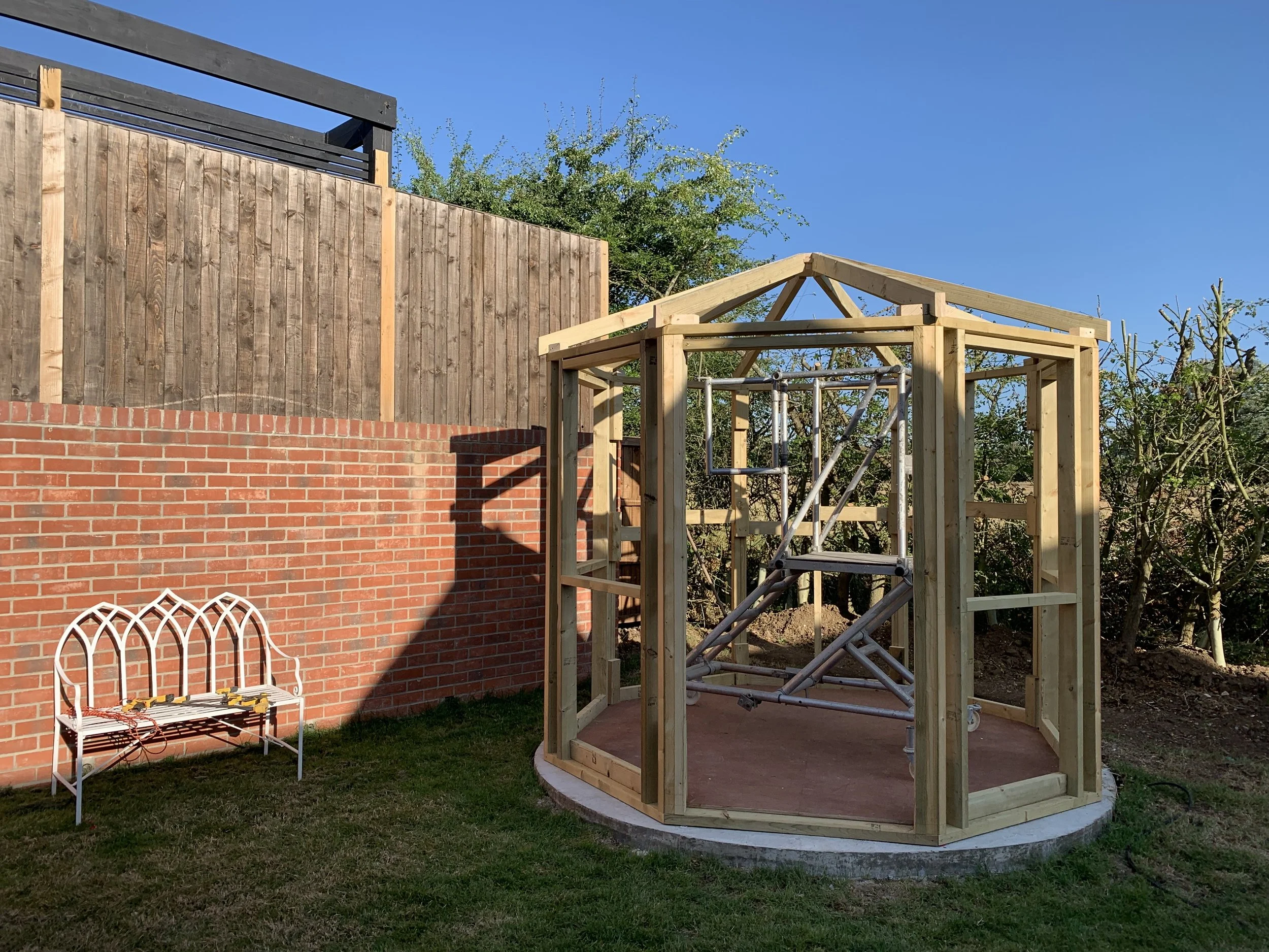 A backyard scene showing a partially built wooden gazebo structure with metal scaffolding inside, a white metal garden bench on the grass, a brick wall, and a wooden fence, with trees and a clear blue sky in the background.