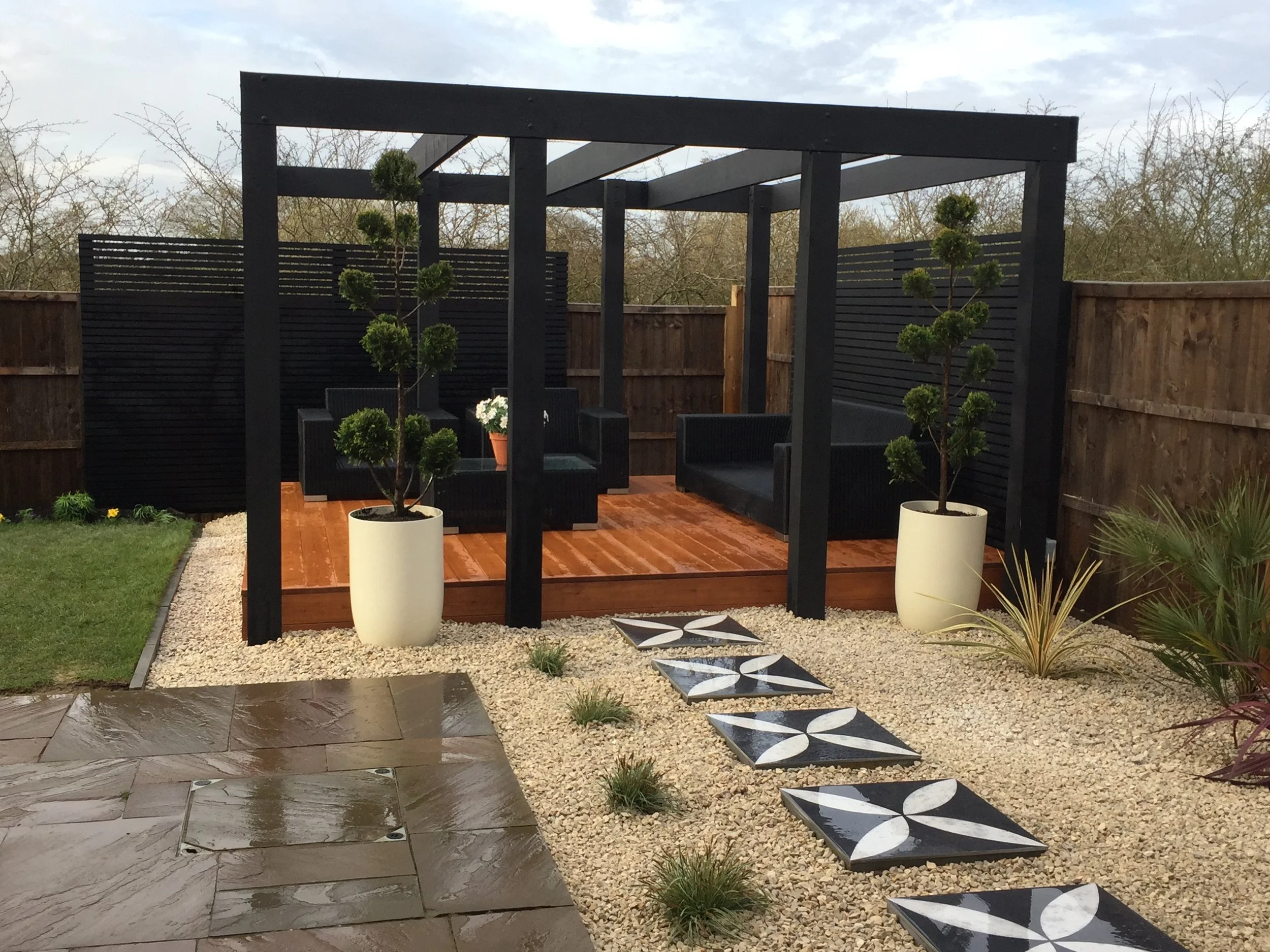 Backyard patio with a wooden deck, black outdoor furniture, two large potted topiary trees, a pathway with patterned tiles, small plants, and a wooden privacy fence with bushes in the background.