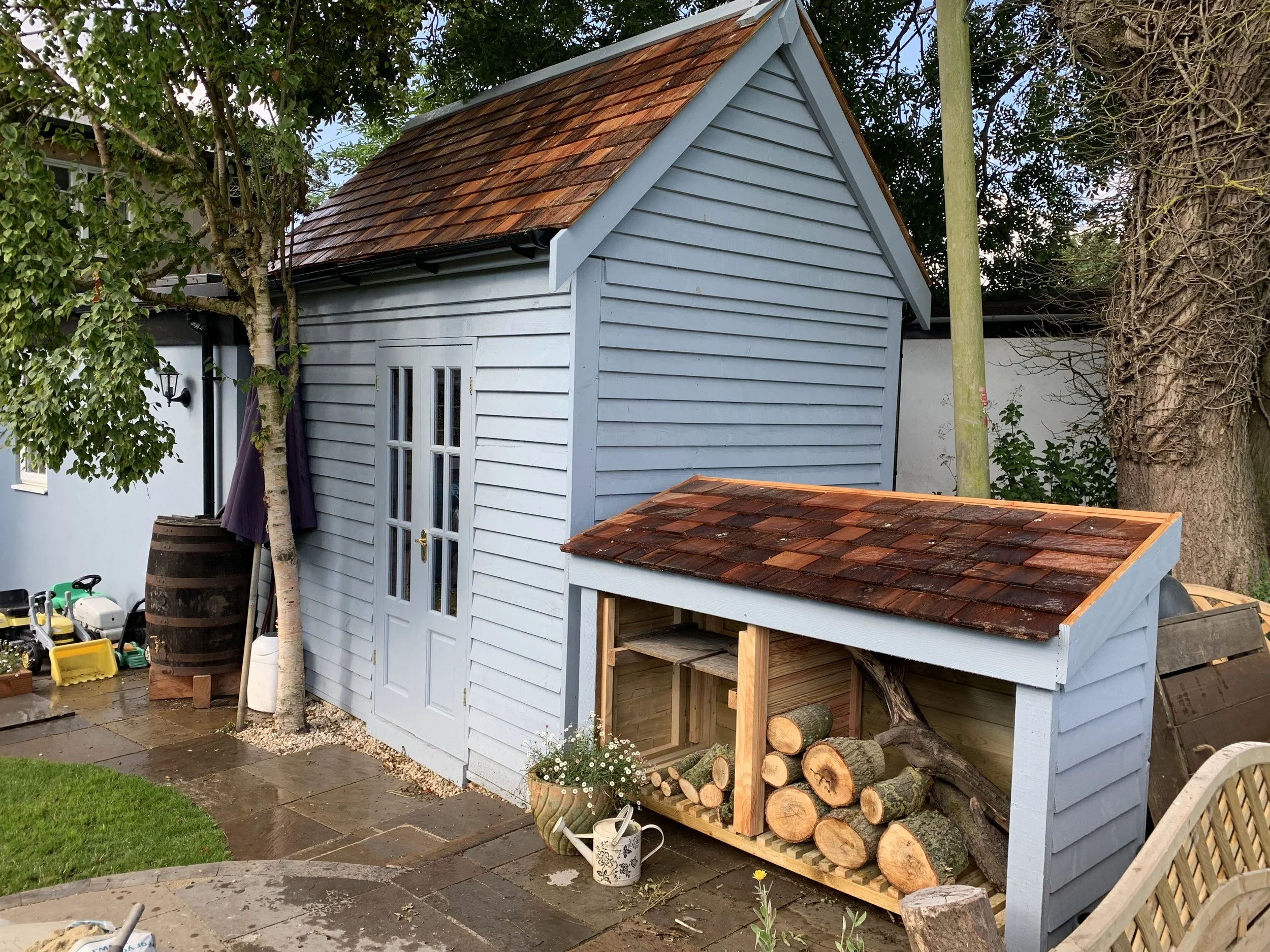 Backyard scene with a light blue wooden shed, log storage with chopped wood, potted plants, and outdoor toys on a wet stone patio after rain.
