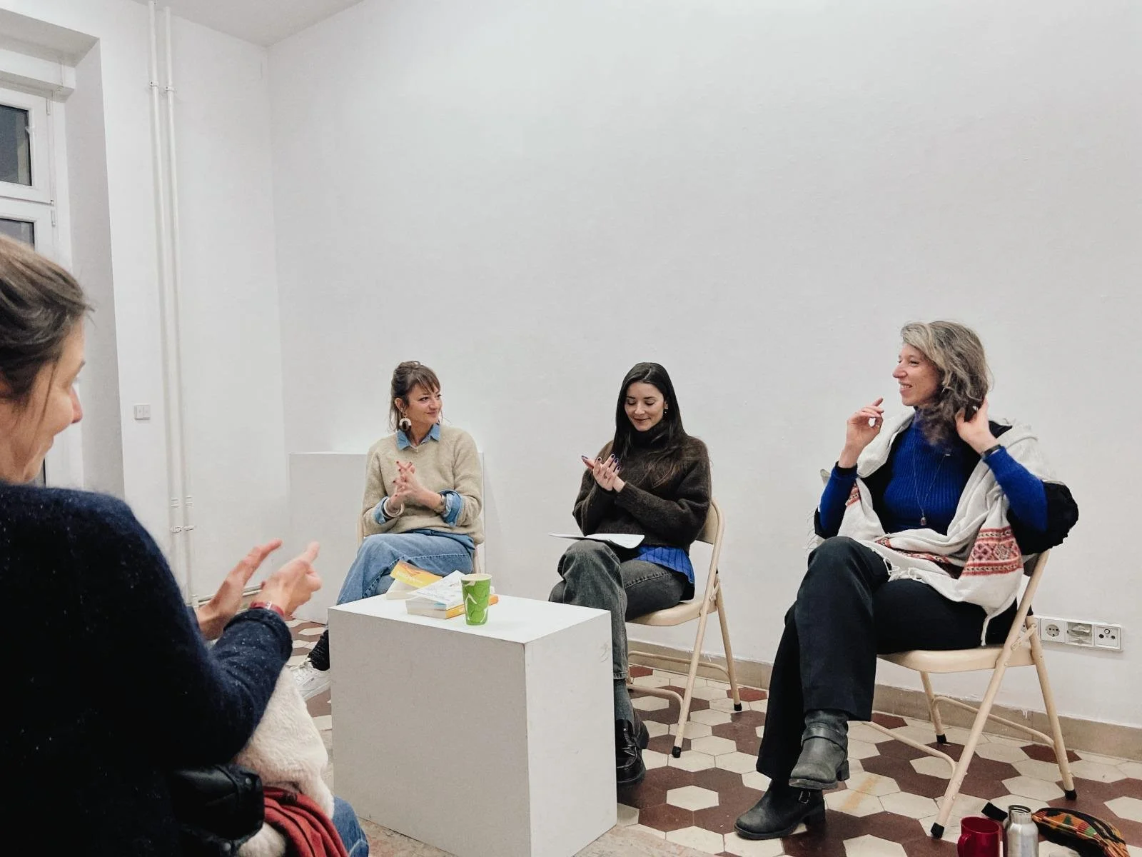 Quatre femmes assises en cercle dans une pièce blanche, en train de discuter. Une femme est en train de parler, d'autres écoutent, deux regardent leur téléphone ou un carnet.